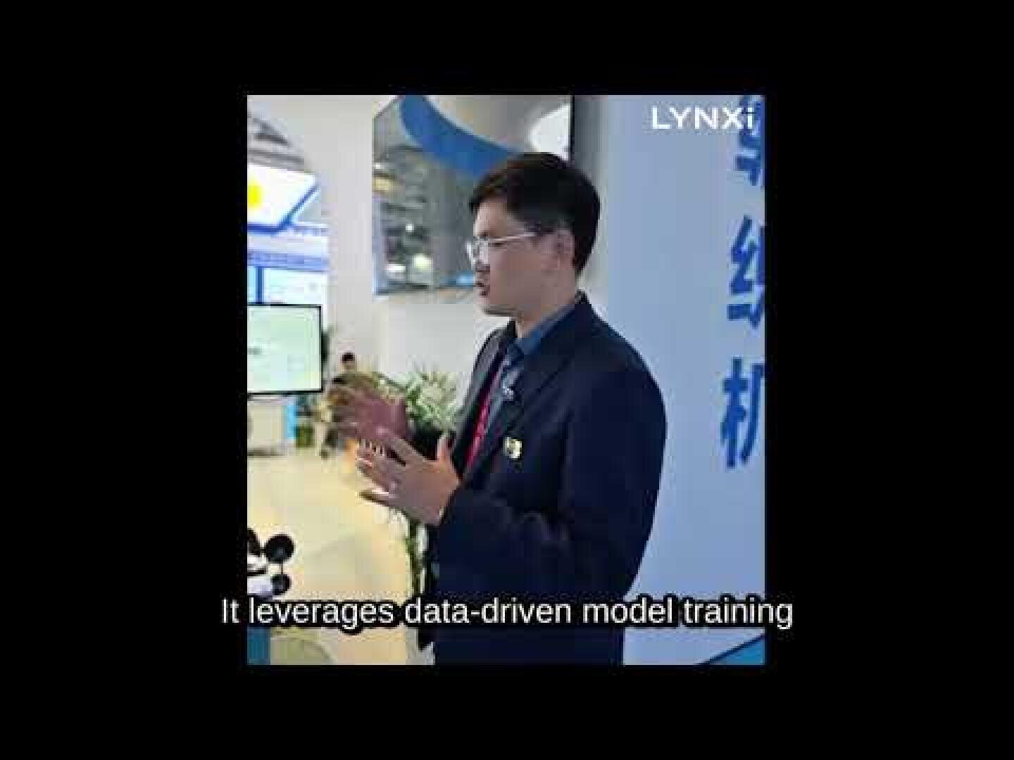 A man in a suit and glasses speaks in front of a display at an event or expo. The word LYNXI is visible in the top right, and subtitles read, It leverages data-driven model training.