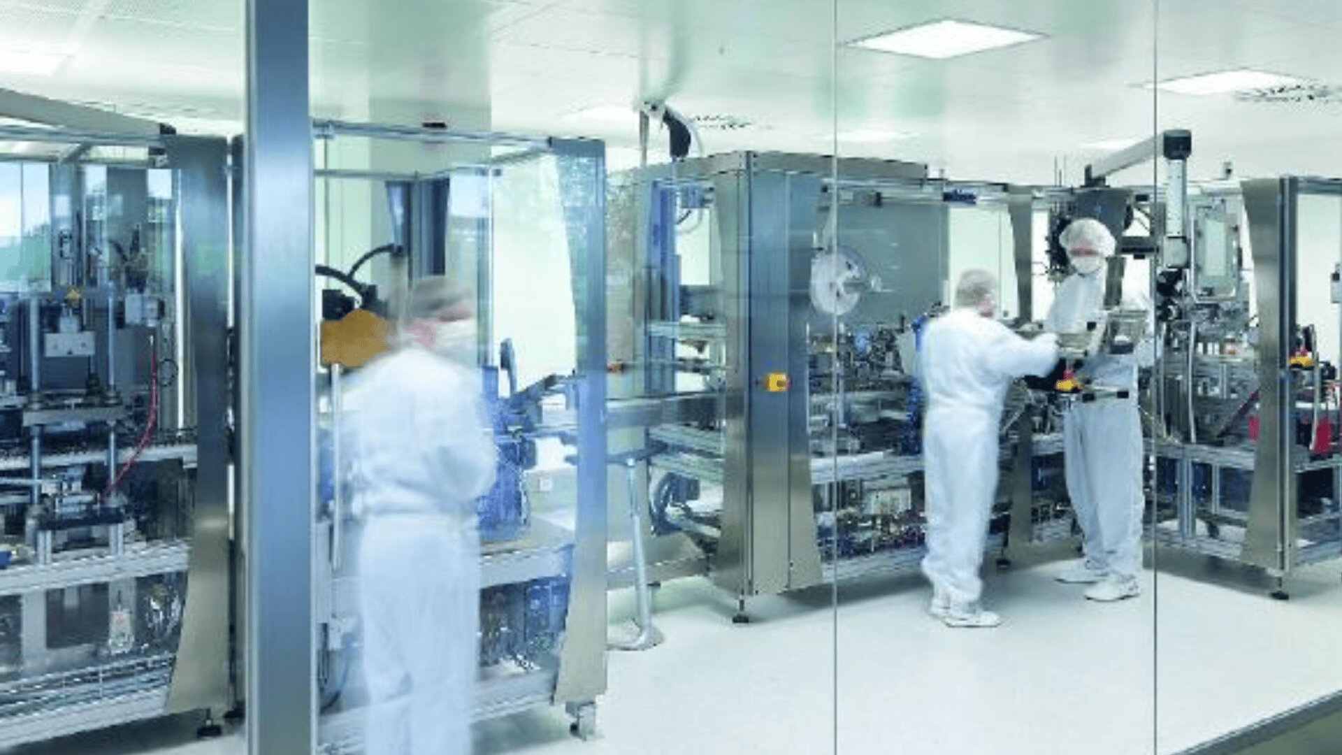 Drei Personen in weißen Schutzanzügen arbeiten mit großen Industriemaschinen in einem sauberen, sterilen Labor oder einer Produktionsanlage, die durch eine Glaswand zu sehen ist. Die Umgebung ist hell und modern.