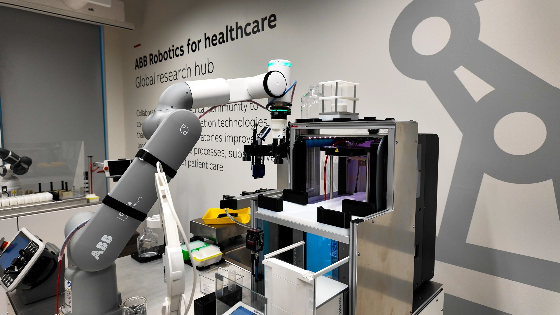 A robotic arm operates laboratory equipment in a modern medical research facility. The words ABB Robotics for Healthcare and the corresponding text are visible on the wall in the background.
