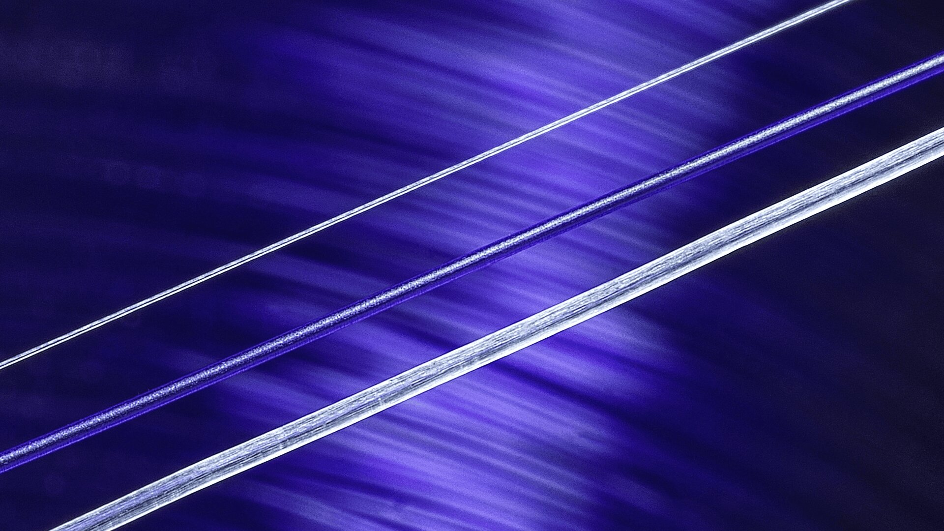 Nahaufnahme von drei diagonalen Metallsaiten vor einem leuchtend blauen und violetten, unscharfen Hintergrund, die an die Saiten eines Musikinstruments erinnern.