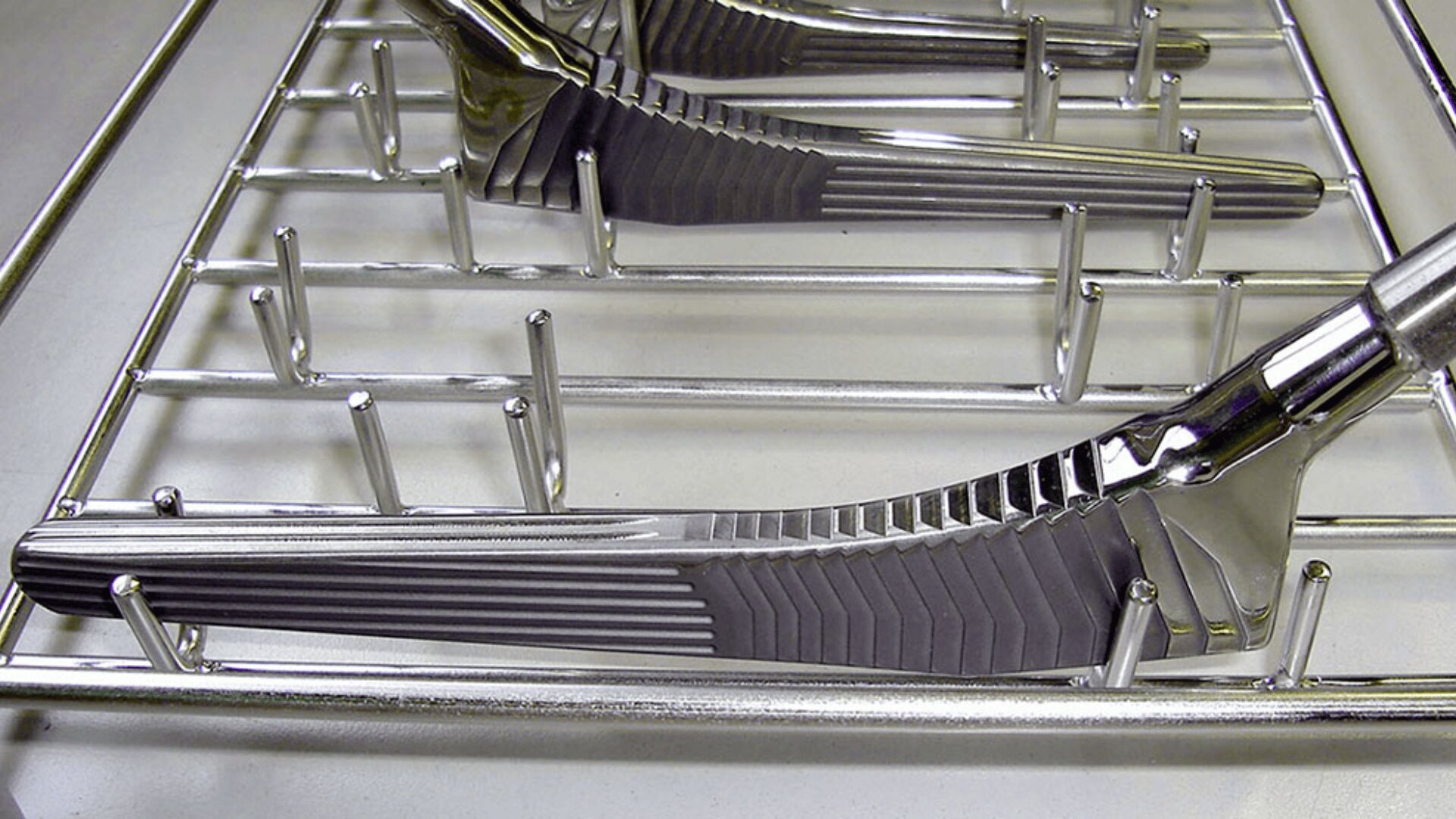 Close-up of several metal butter knives arranged neatly in the utensil rack of a dishwasher, with the handles facing outward and the blades secured between metal prongs.