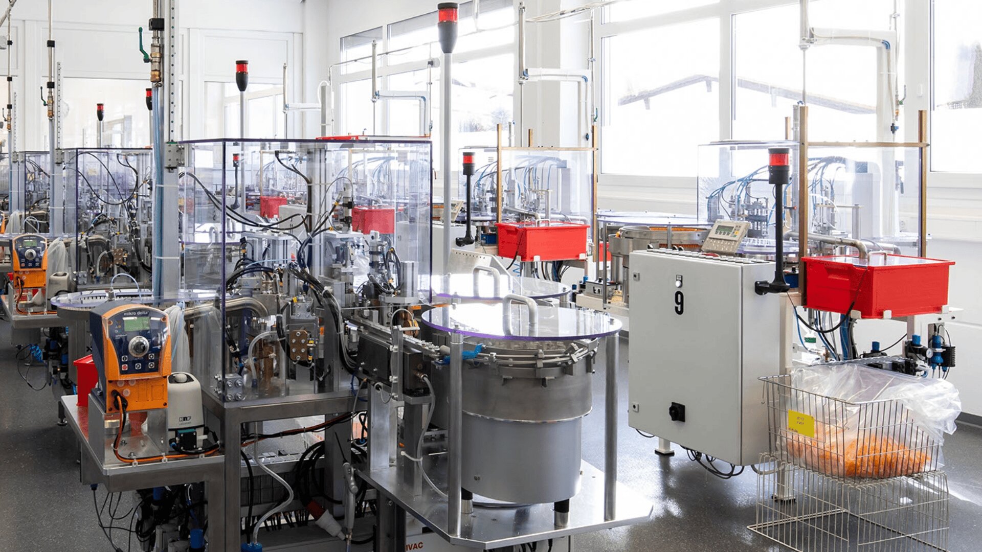 Eine hell erleuchtete Fabrik mit automatisierten Industriemaschinen und -anlagen zur Materialverarbeitung, umgeben von transparenten Sicherheitsbarrieren und Bedienfeldern. Große Fenster sorgen für natürliches Licht im sauberen, organisierten Arbeitsbereich.