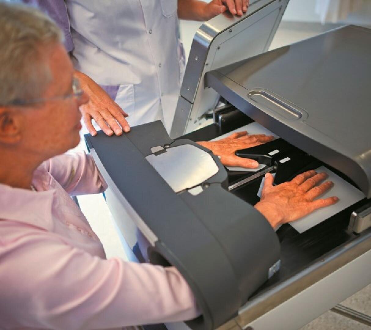 An older adult places both hands into a medical scanner, assisted by a healthcare professional, likely for diagnostic imaging or bone density testing.