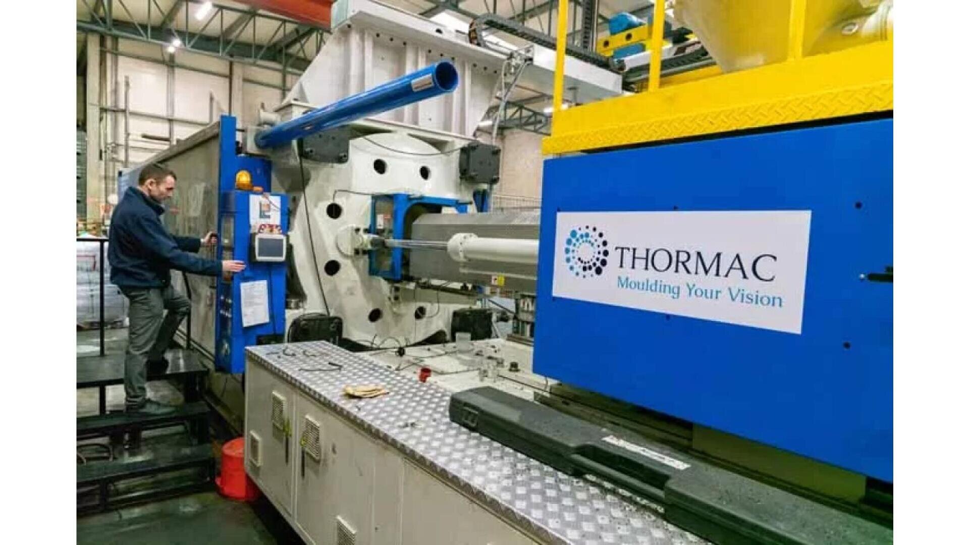Eine Person bedient in einer Fabrik eine große blau-weiße Spritzgussmaschine. An der Maschine hängt ein Schild mit der Aufschrift „THORMAC Moulding Your Vision“. Verschiedene Geräte und Werkzeuge sind rund um den Arbeitsplatz zu sehen.