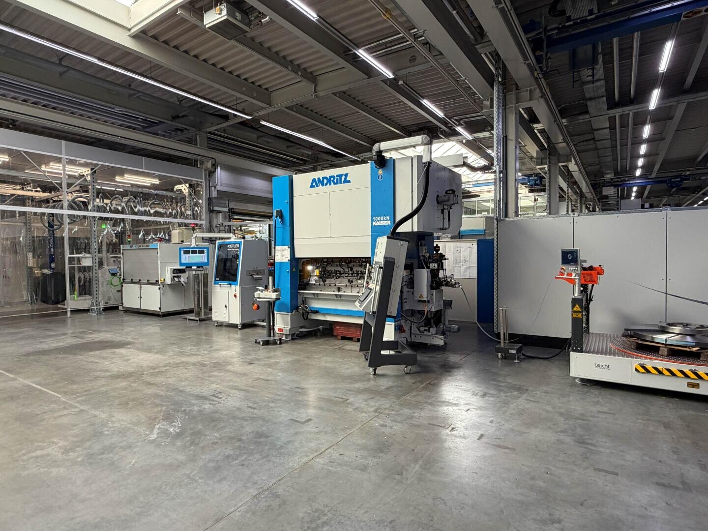 Eine geräumige Industrieanlage mit einer großen blau-weißen ANDRITZ-Maschine, verschiedenen Schalttafeln, Geräten und automatisierten Maschinen auf einem polierten Betonboden unter hellen Oberlichtern.