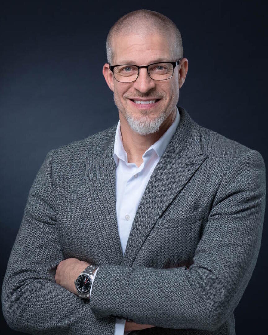 Ein lächelnder Mann mit Brille, kurzem Bart und kurz geschnittenem Haar trägt einen grauen strukturierten Blazer über einem weißen Hemd. Er steht mit verschränkten Armen vor einem dunklen Hintergrund.