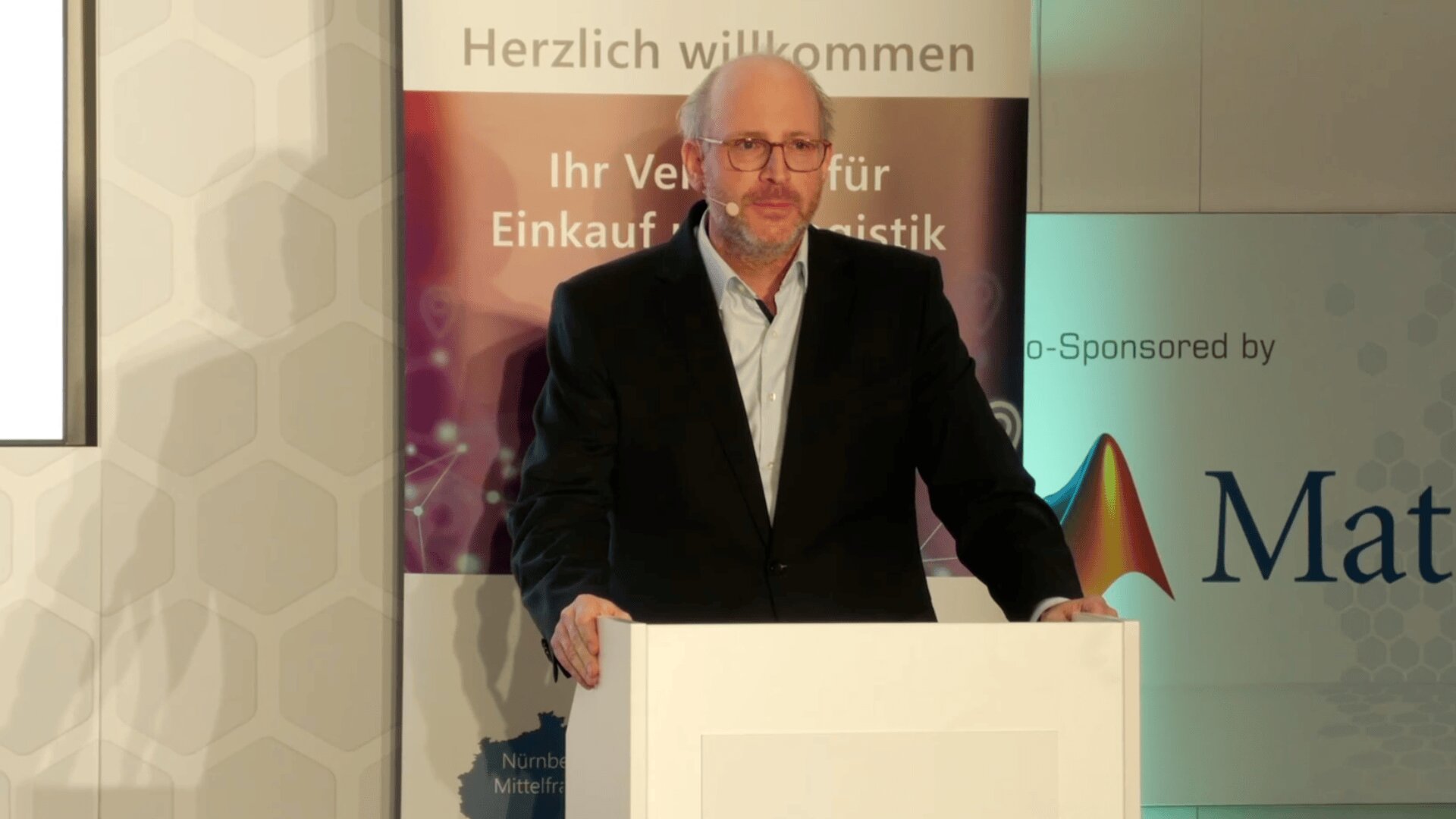 Ein Mann in einem schwarzen Anzug und mit Brille steht an einem weißen Podium und spricht auf einer Veranstaltung. Hinter ihm steht ein Schild mit der Aufschrift "Herzlich willkommen" und ein Banner mit Logos und Sponsorenangaben.
