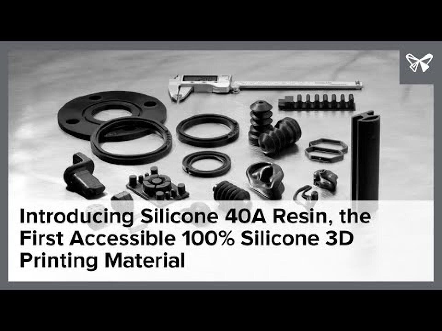 A variety of black 3D-printed silicone parts and tools, including rings, boots, and gears, are arranged on a table. Overlaid text reads: “Introducing Silicone 40A Resin, the First Accessible 100% Silicone 3D Printing Material.”.