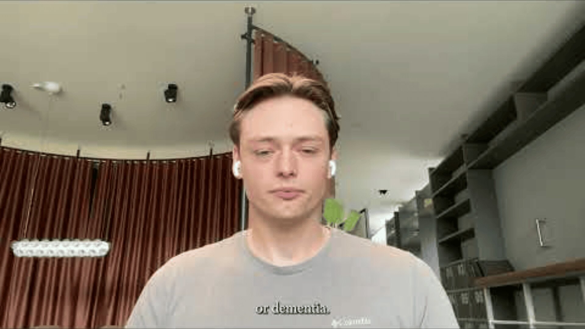 A young man with light brown hair wearing a gray shirt and wireless earbuds sits indoors with brown curtains and shelves in the background. Subtitles at the bottom read, or dementia.