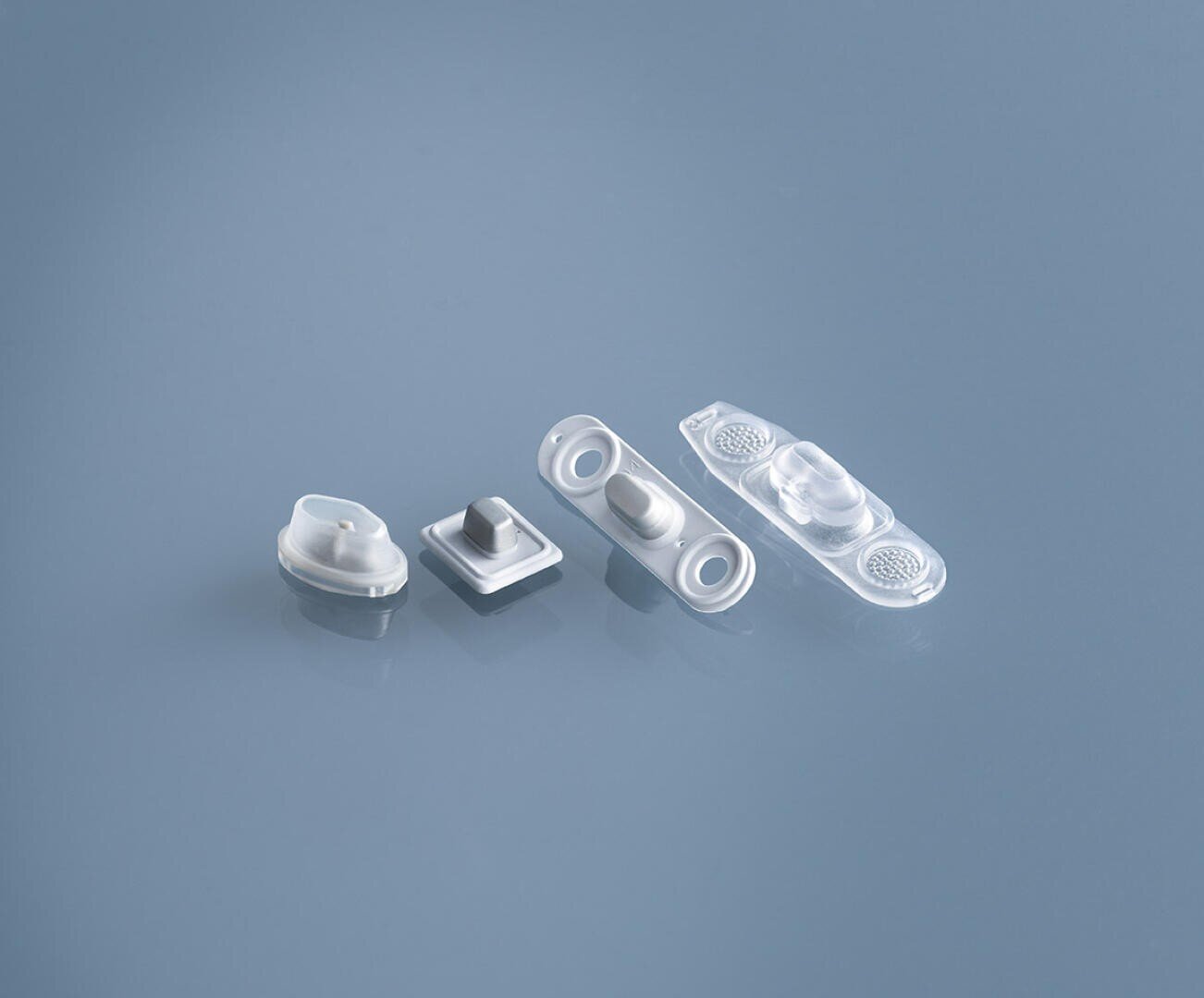 Four small, white and transparent plastic medical or laboratory components are arranged in a row on a smooth, light blue surface. Each piece has a distinct shape and design.