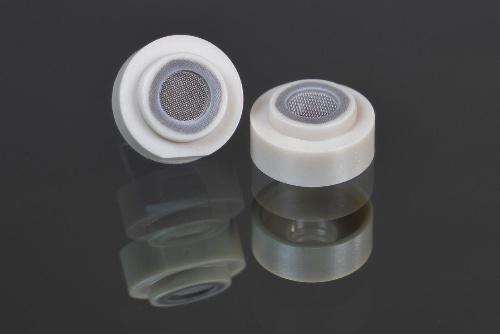 Two small white circular plastic components with mesh screens in the center are displayed on a reflective black surface, showing their reflection below them.