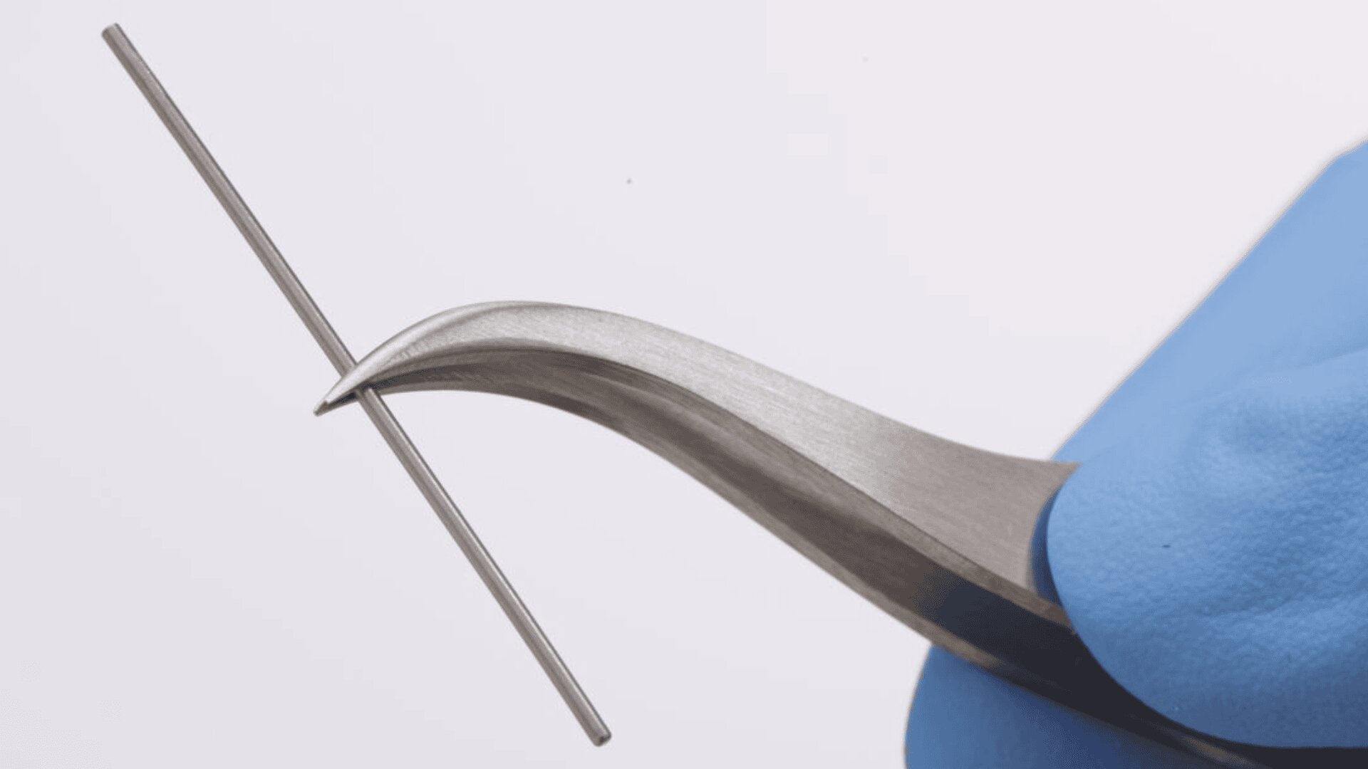 Eine behandschuhte Hand hält eine gebogene Pinzette und greift damit die Mitte eines dünnen, geraden Metallstabs vor einem schlichten, hellen Hintergrund.