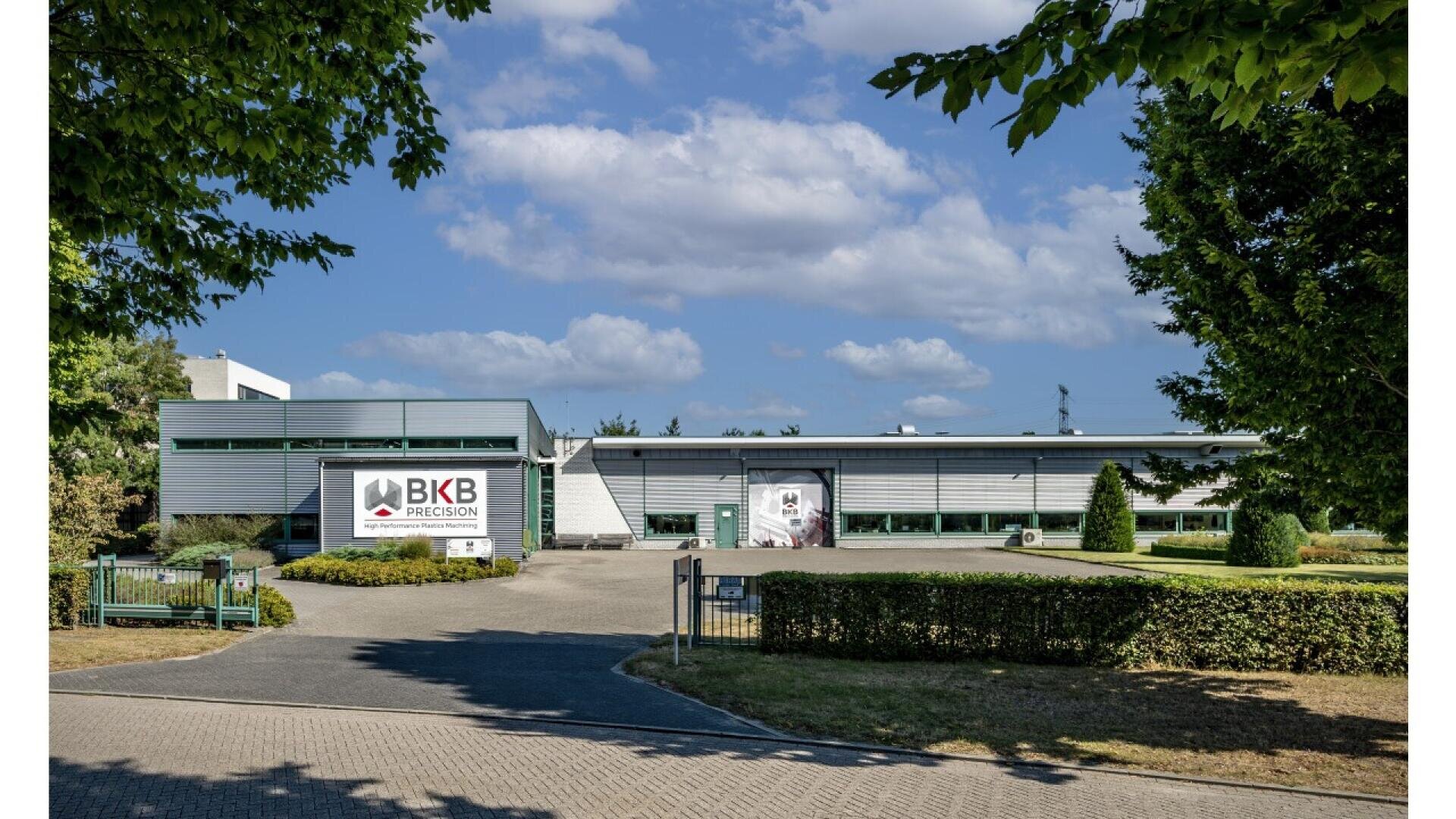 A modern industrial building with a sign reading BKB Precision on the facade, surrounded by neatly trimmed bushes, trees, a small lawn, and a driveway under a partly cloudy sky.