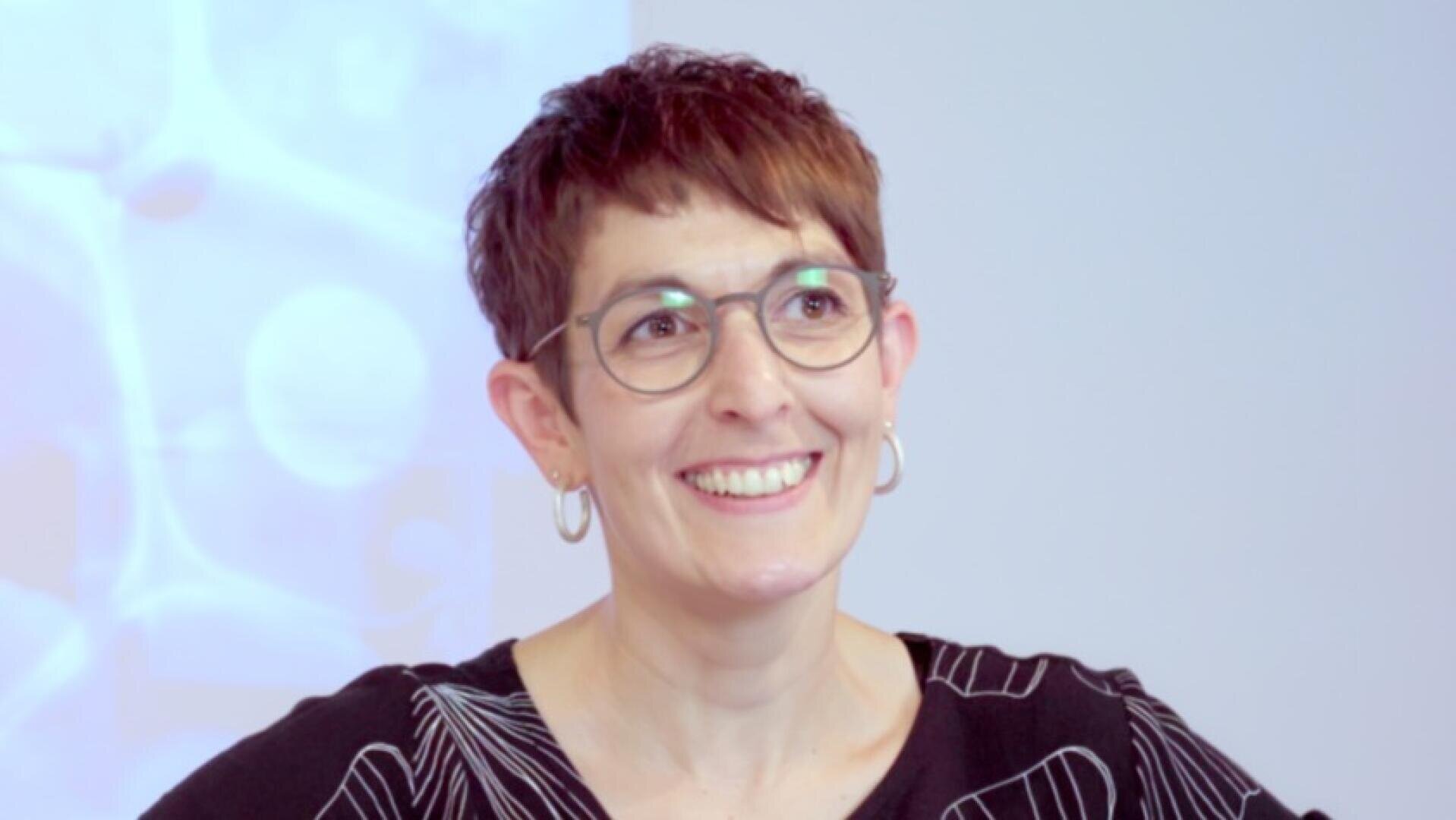 Eine lächelnde Frau mit kurzen braunen Haaren, die eine Brille, Ohrringe und ein schwarzes Oberteil mit weißen Linienmustern trägt, steht vor einem hellblauen Hintergrund mit abstrakten Formen.