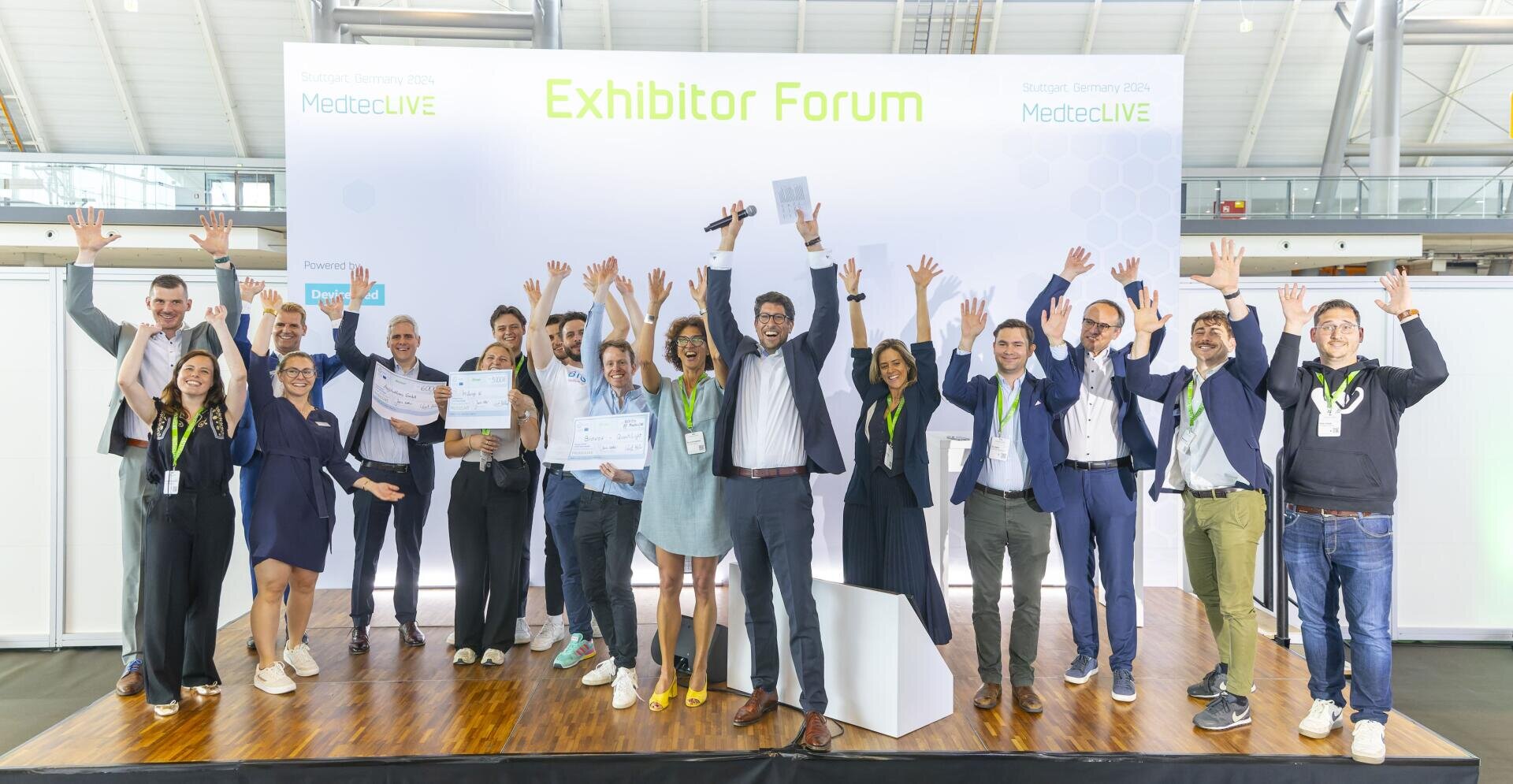 A group of people on the stage of the MedtecLIVE exhibitor forum joyfully raise their hands in celebration. Some are holding certificates, one person is holding a microphone. A large Exhibitor Forum sign can be seen above them.