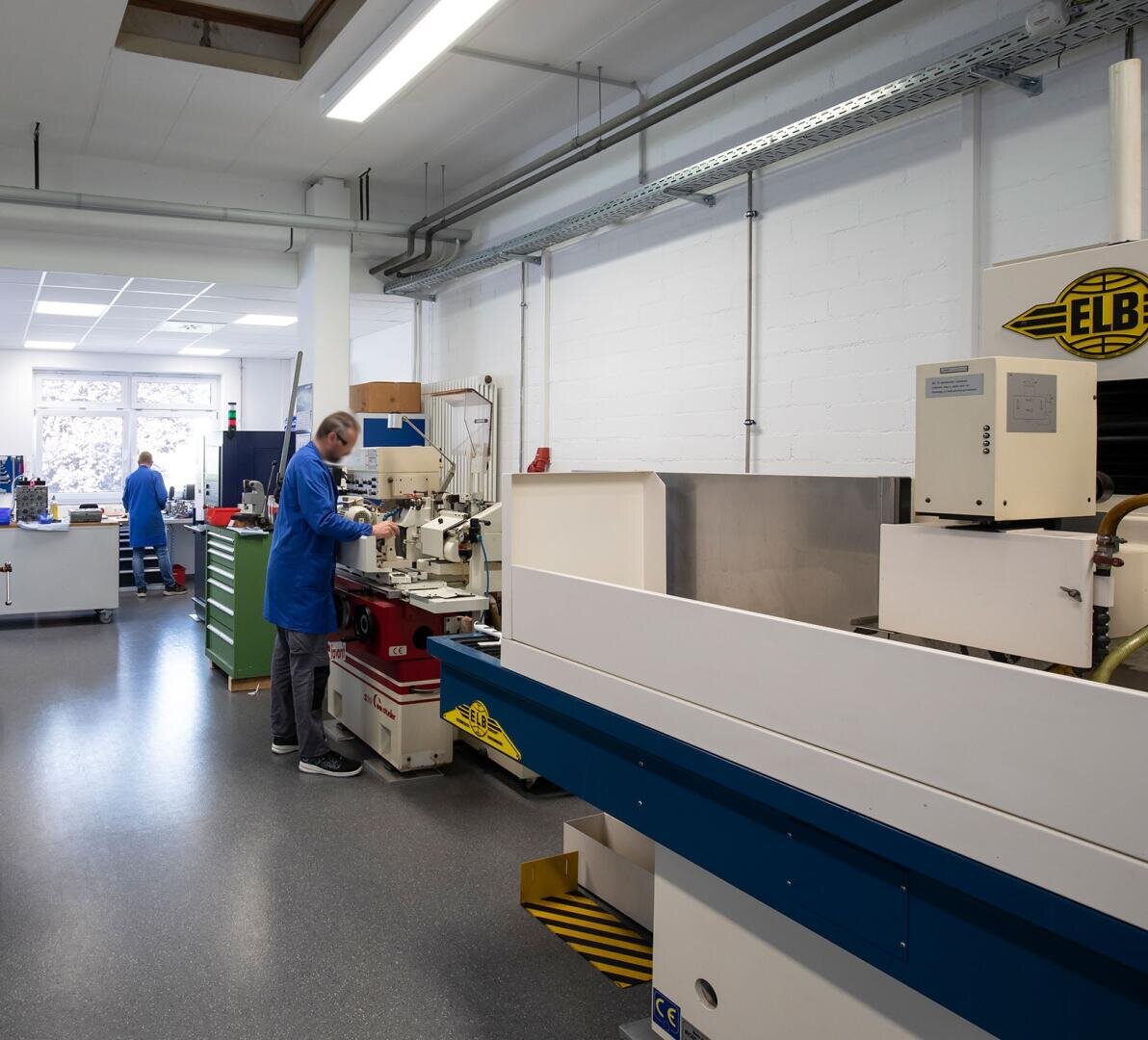 Ein Mann im blauen Laborkittel bedient Maschinen in einer sauberen, organisierten Werkstatt mit Industrieanlagen und Schränken. Im Hintergrund arbeitet eine weitere Person in der Nähe großer Fenster.