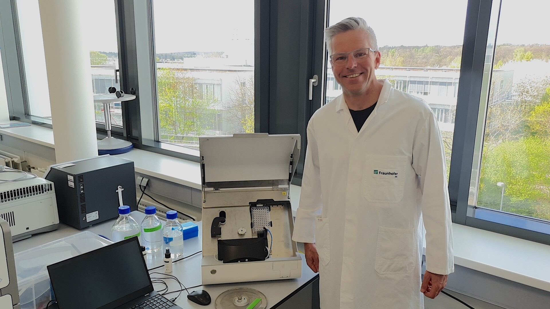 Andreas Traube in a white lab coat stands next to scientific equipment on a lab bench by large windows, with bottles, a laptop, and other lab items visible in a bright laboratory setting.