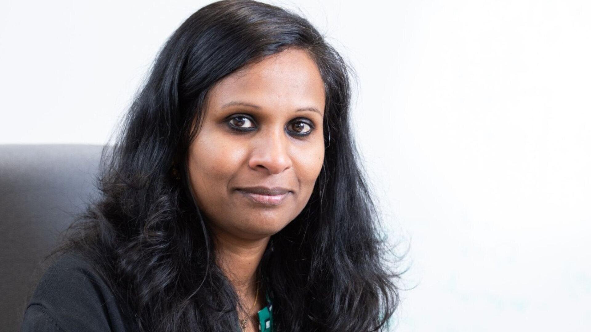A woman with long, dark hair and brown skin sits and smiles softly at the camera. She is wearing a dark top and is pictured against a plain, light background.
