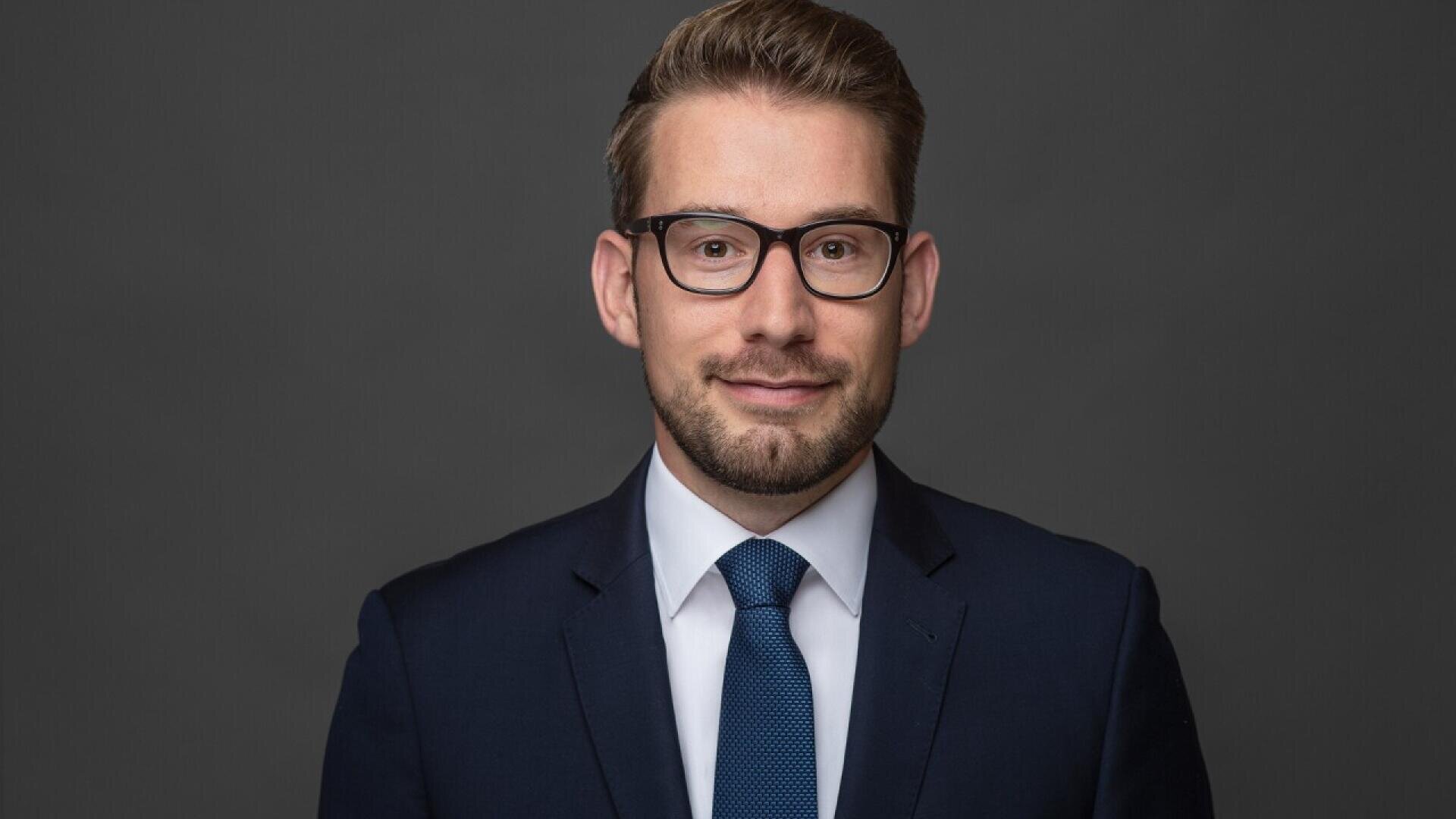 A man in a dark suit, white shirt, and blue tie stands against a plain dark background. He has short brown hair, a trimmed beard, wears black-rimmed glasses, and is smiling slightly.