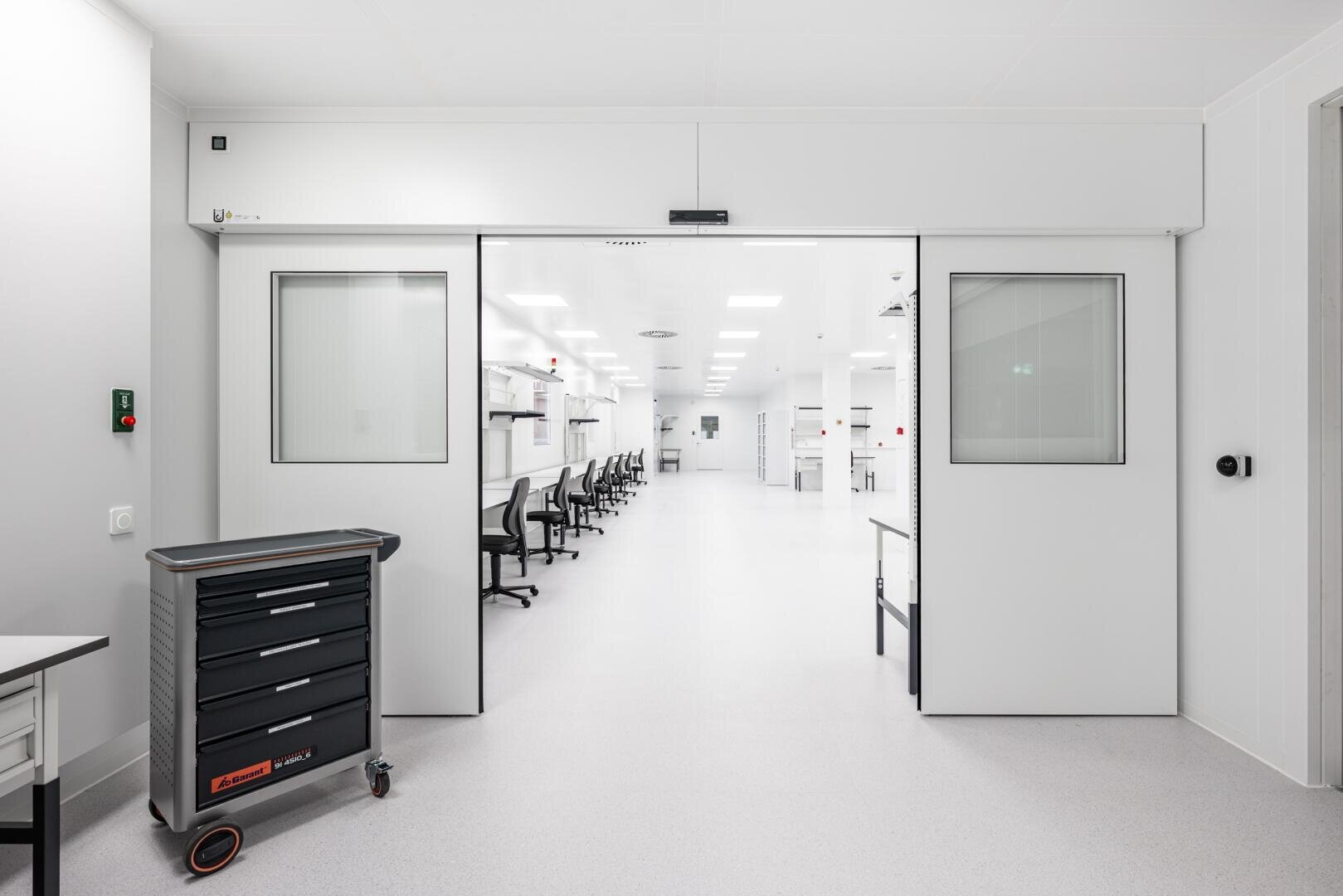 A bright, modern laboratory with white walls and floors, black wheelchairs at the workstations and a tool trolley next to wide automatic sliding doors. The room is clean, tidy and well lit with skylights.