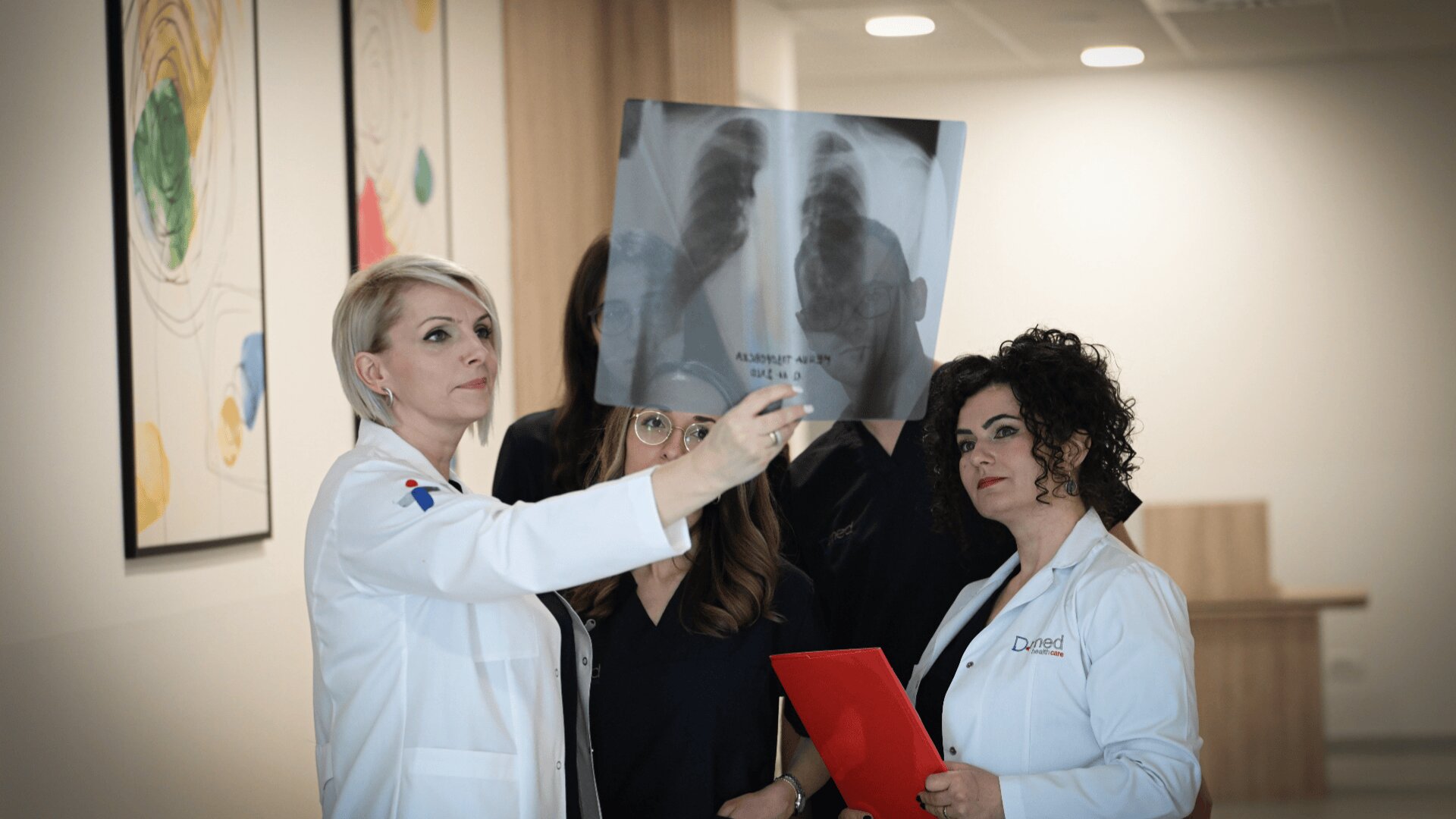 Three doctors in white coats examine an X-ray film together in a modern medical office. One doctor holds the X-ray up, while another holds a red folder. Artwork and furniture are visible in the background.
