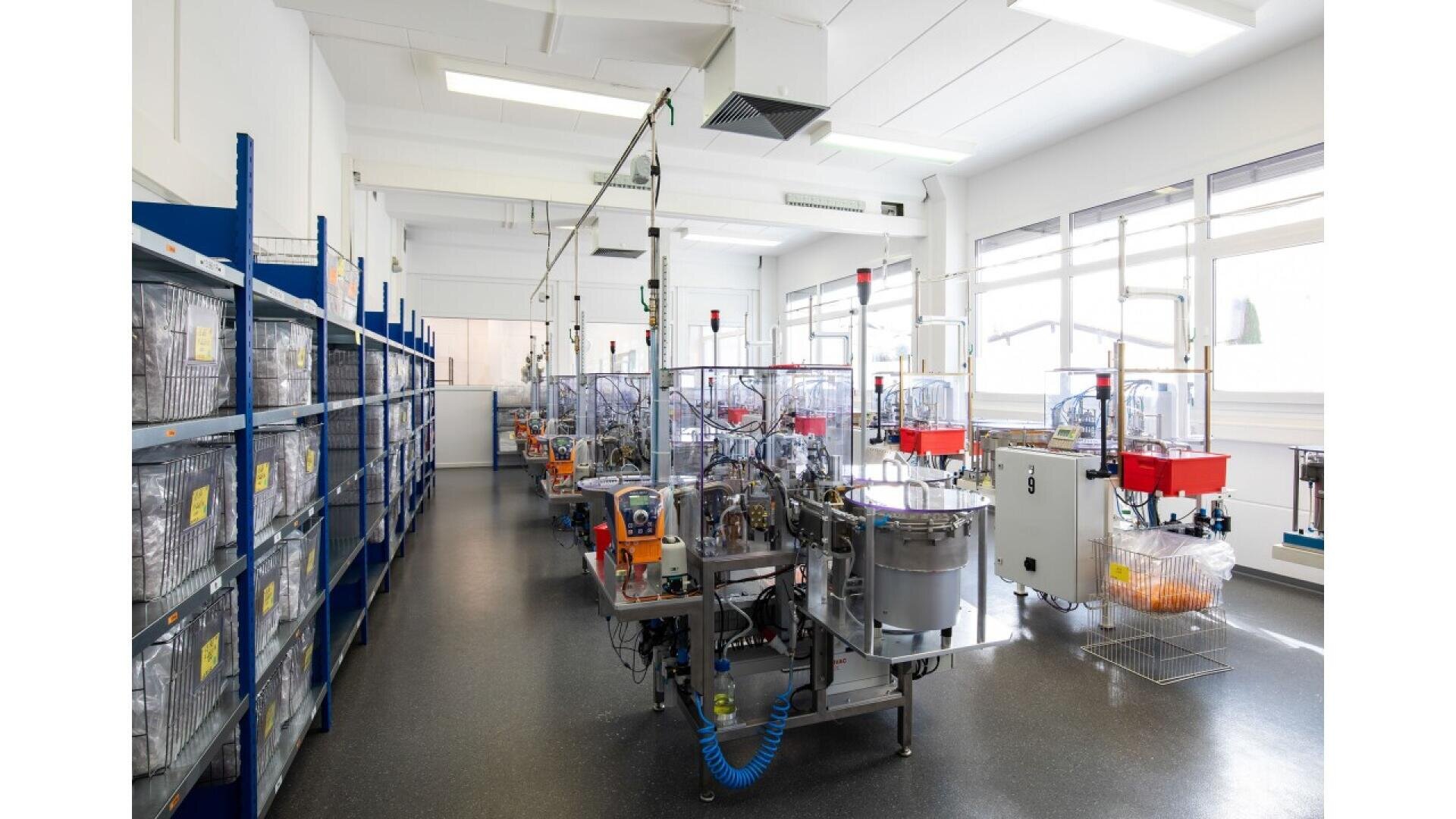 A clean, brightly lit laboratory with rows of automated equipment and clear enclosures, shelves filled with containers on the left, and large windows letting in natural light on the right.