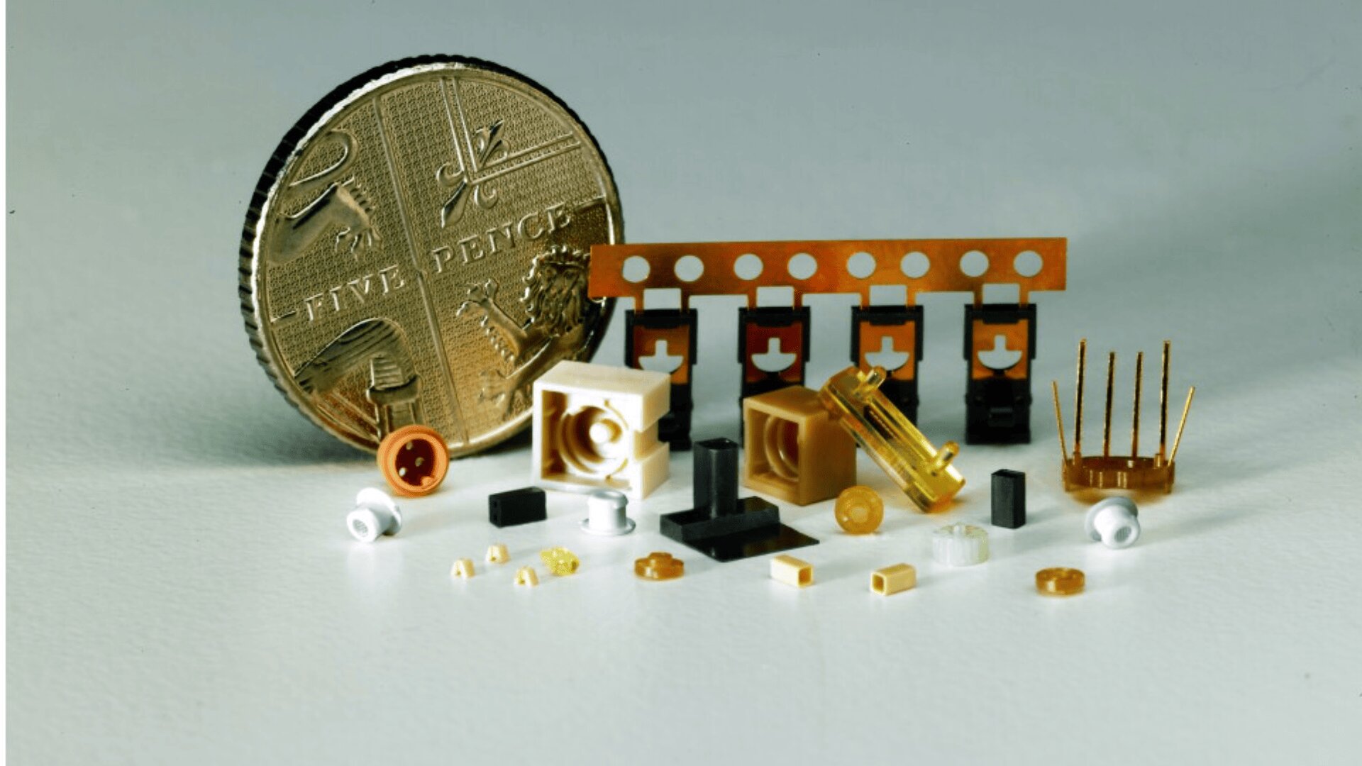 Eine britische Fünf-Pence-Münze wird neben mehreren winzigen elektronischen und mechanischen Bauteilen platziert, um deren geringe Größe hervorzuheben. Die Bauteile haben verschiedene Formen und Farben, darunter Gold, Schwarz, Weiß und transparente Teile.