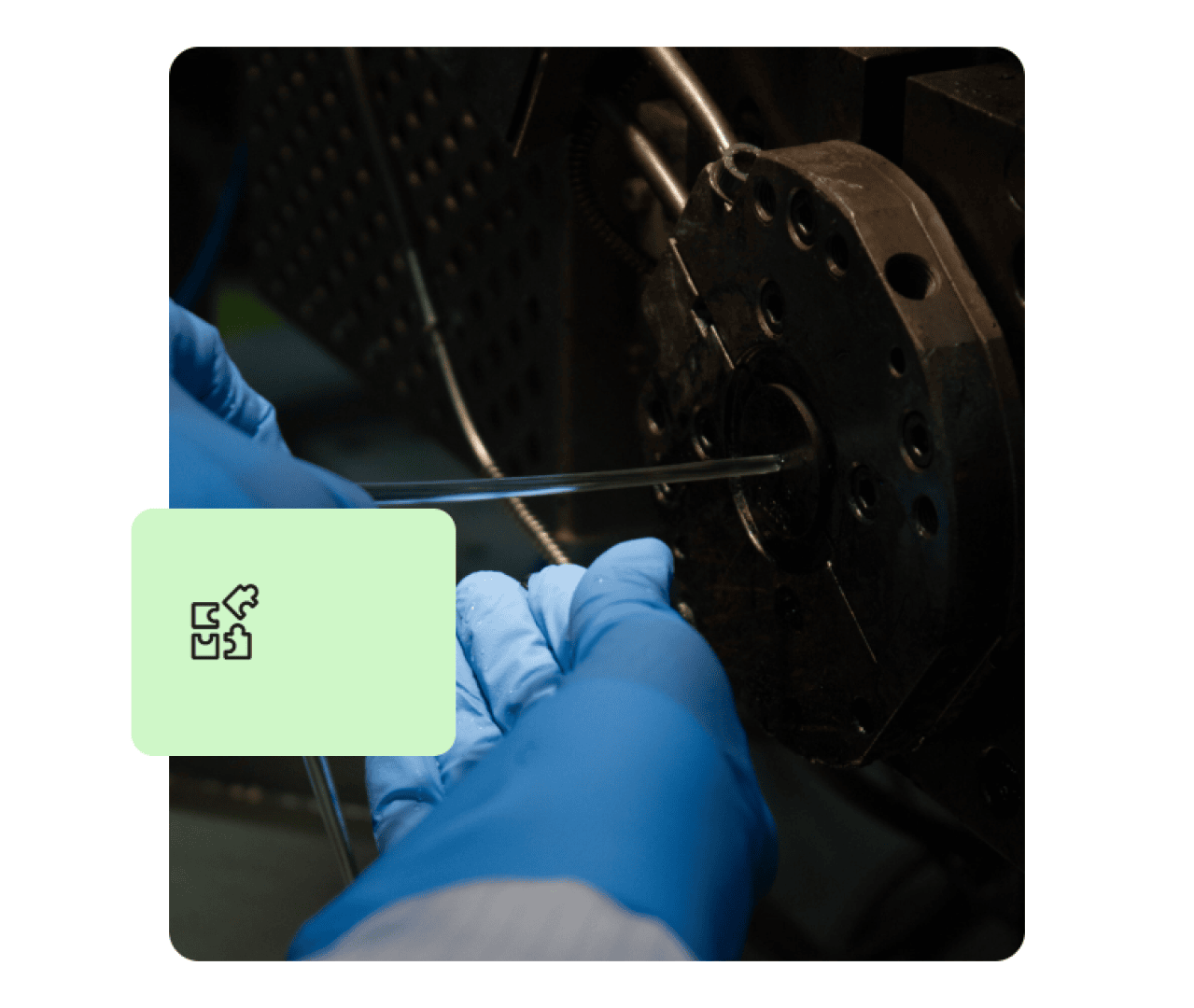 A person wearing blue gloves inserts a clear tube into a metal machine part. A pale green box with an abstract icon partially overlays the image.