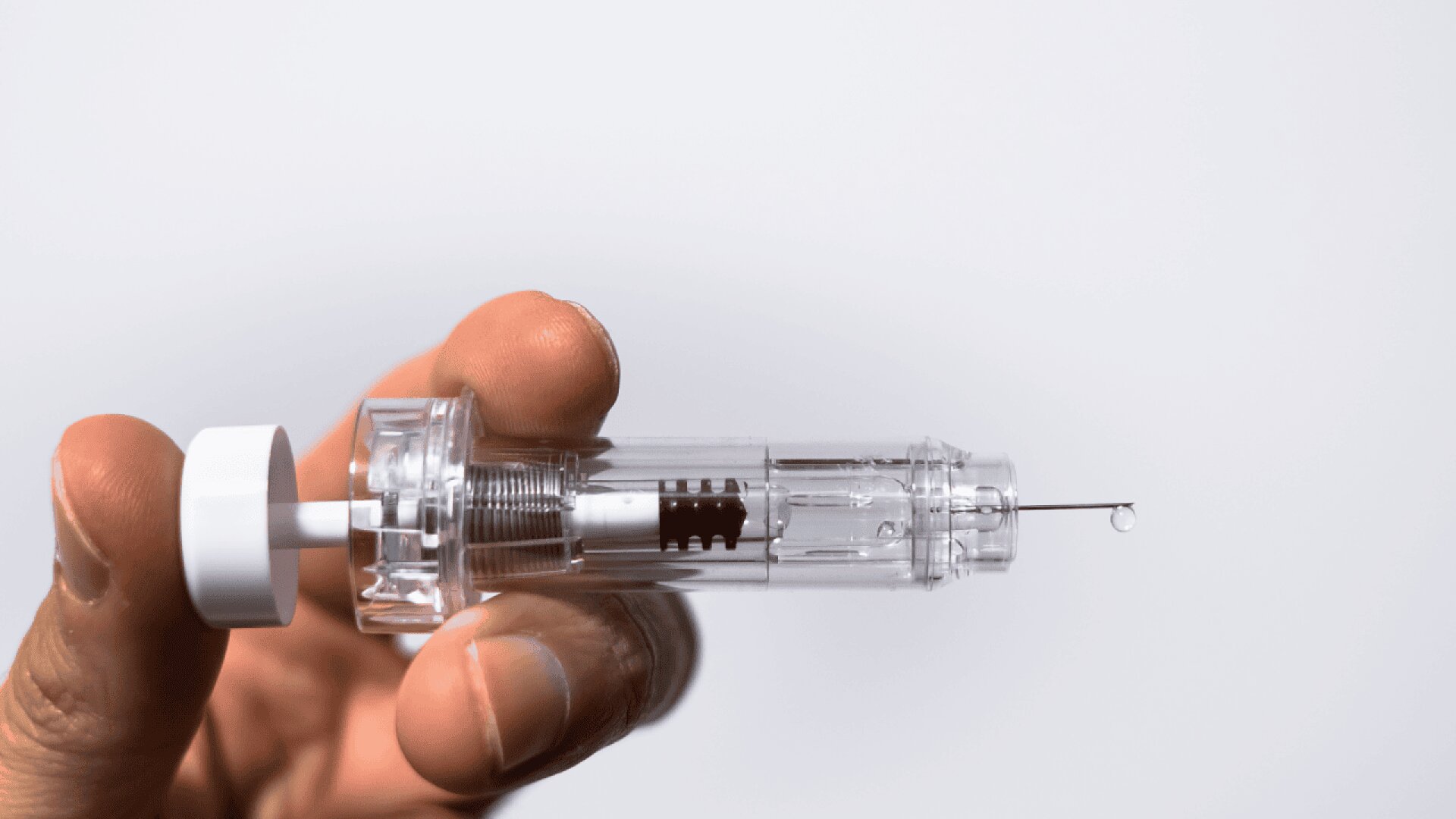 A close-up of a hand holding a clear, plastic medical syringe with a visible needle and a droplet of liquid at the tip, against a plain white background.