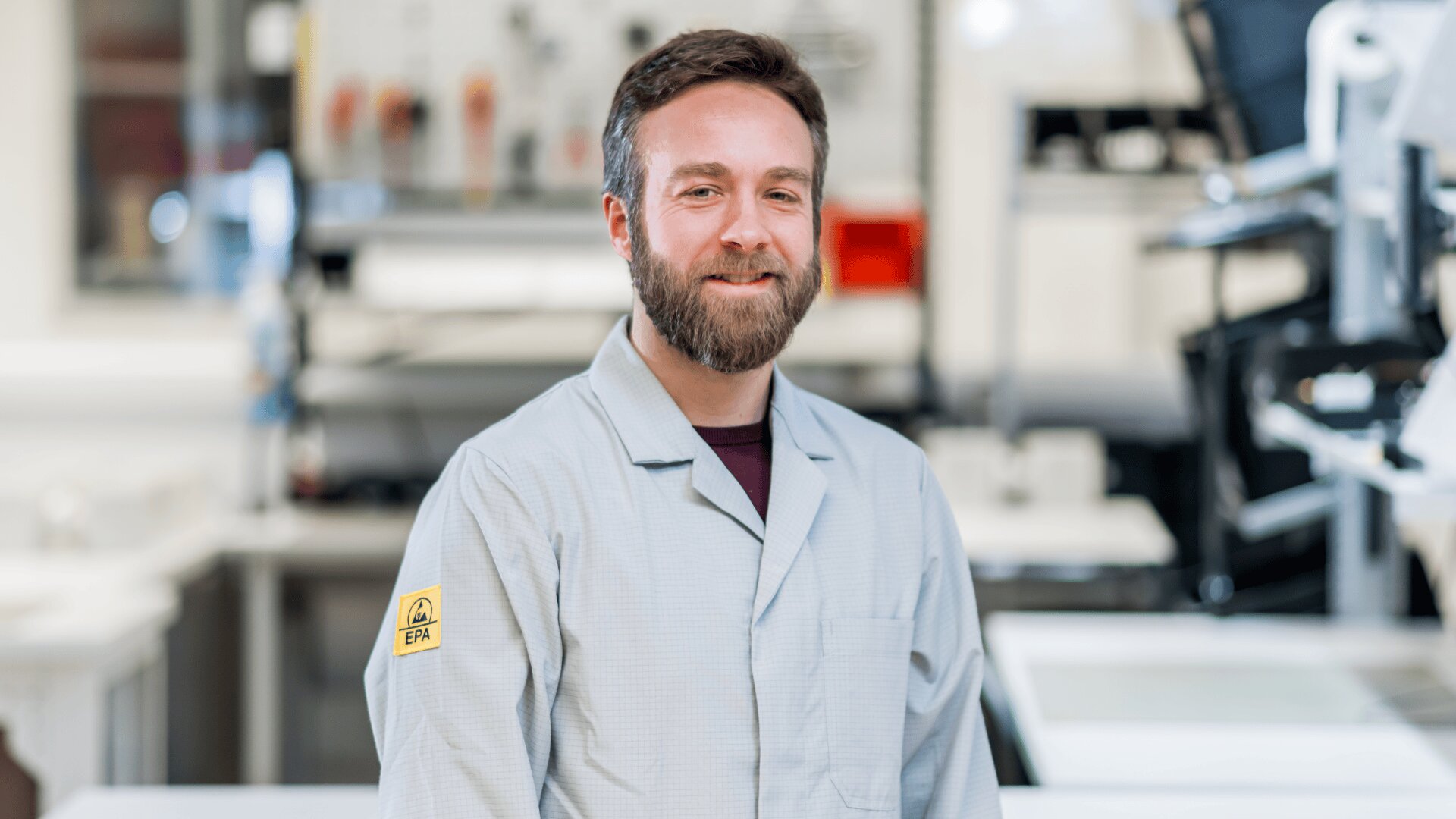 Ein Mann mit Bart, der einen hellgrauen Laborkittel mit einem ESD-Symbol (elektrostatische Entladung) auf dem Ärmel trägt, steht lächelnd in einer Laborumgebung mit Geräten im Hintergrund.