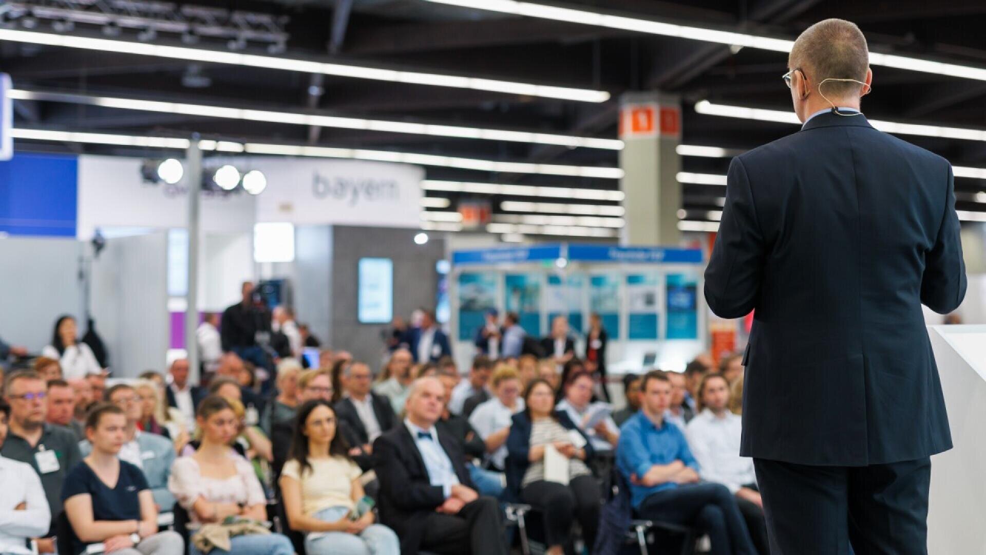 A man in a suit, seen from behind, speaks to a large audience seated in rows at a conference or seminar in a brightly lit convention hall.