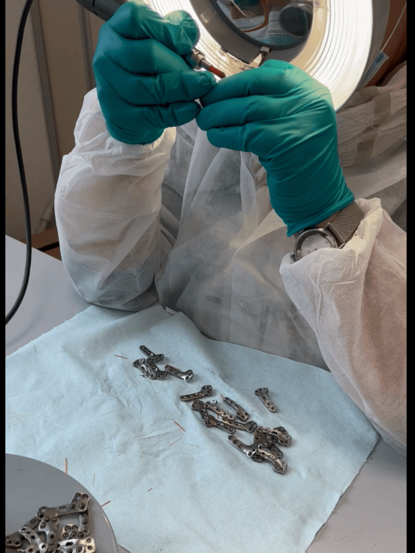 A person wearing teal gloves and a white lab coat examines a small metal object under a magnifying lamp, with several similar metal parts laid out on a blue cloth on the table.