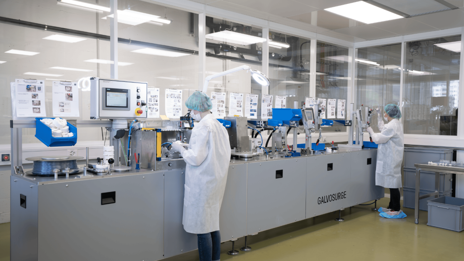 Zwei Mitarbeiter in weißen Laborkitteln, Haarnetzen und Schuhüberzügen bedienen Maschinen in einem sauberen, modernen Labor oder einer Produktionsstätte mit großen Fenstern und technischen Geräten.