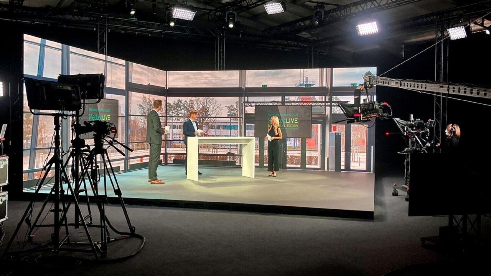 Three people stand and talk at a white table in a brightly lit TV studio with multiple cameras, lighting equipment, and large digital screens displaying a cityscape in the background.
