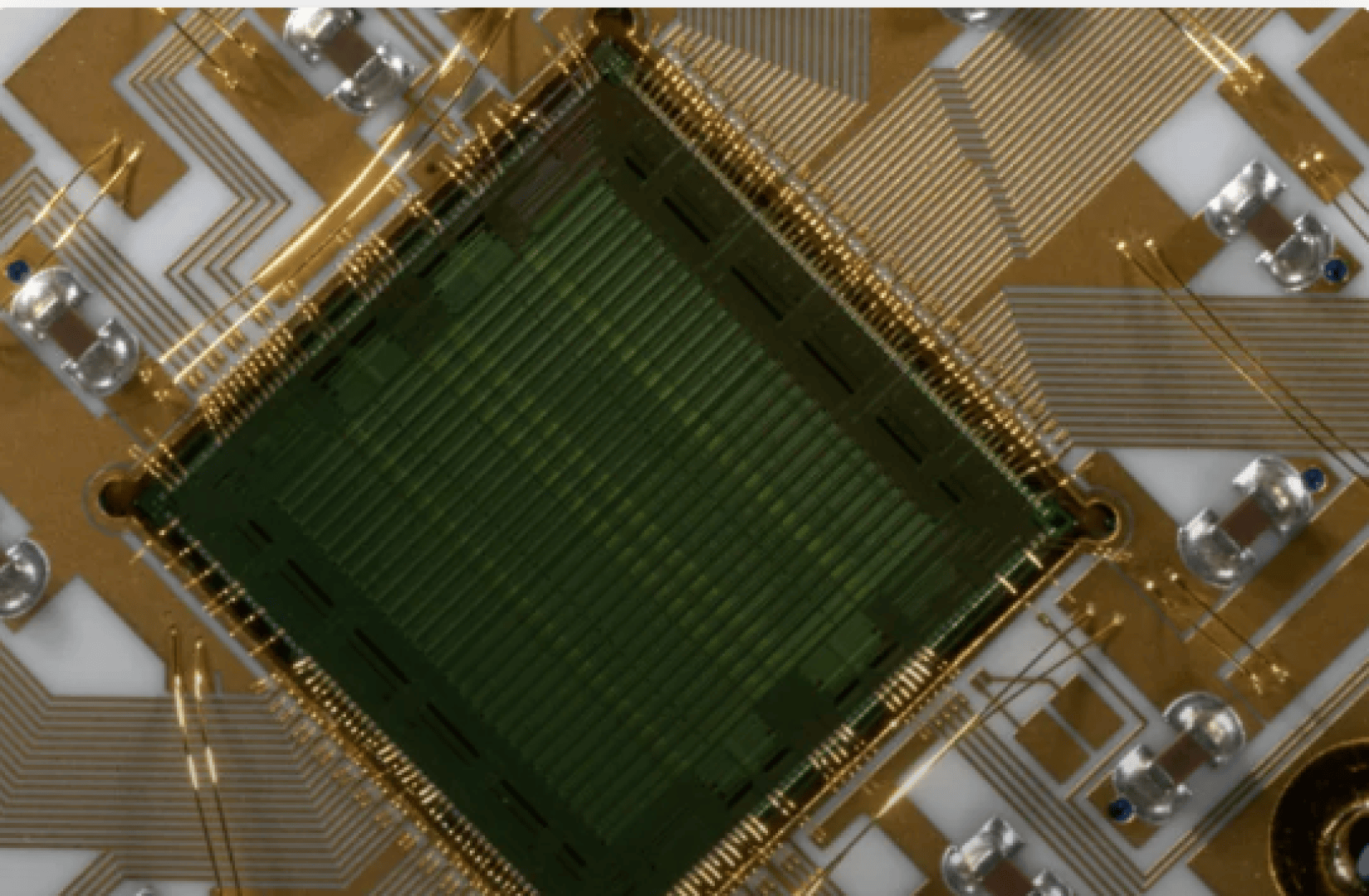 Close-up view of a computer chip or integrated circuit with gold wiring, metallic connectors, and a green central component arranged in a square pattern on a circuit board.
