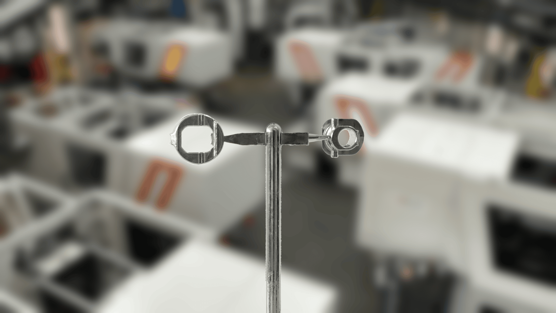Close-up of a metal tool with two differently sized square and round openings, held upright in front of a blurred background of white industrial machinery.