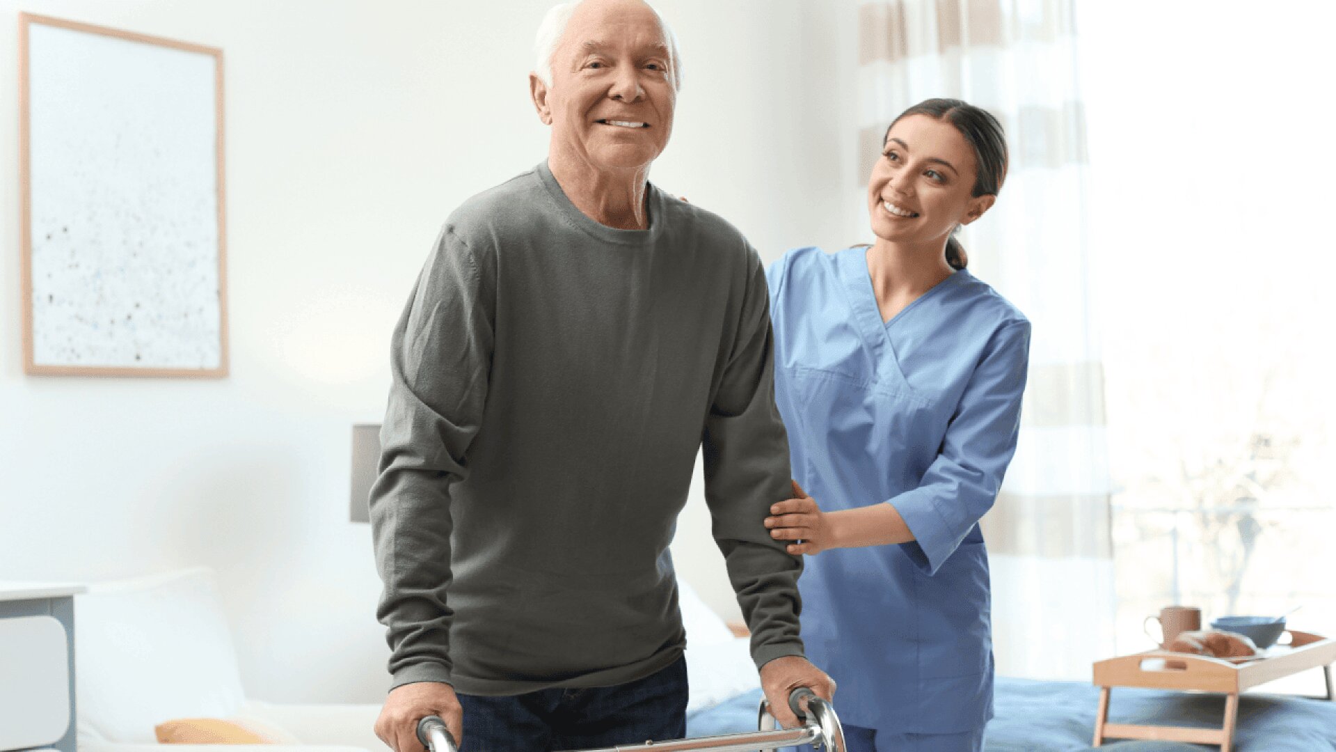An elderly man using a walker smiles while a young female caregiver in blue scrubs stands beside him, offering support in a bright, cozy room.