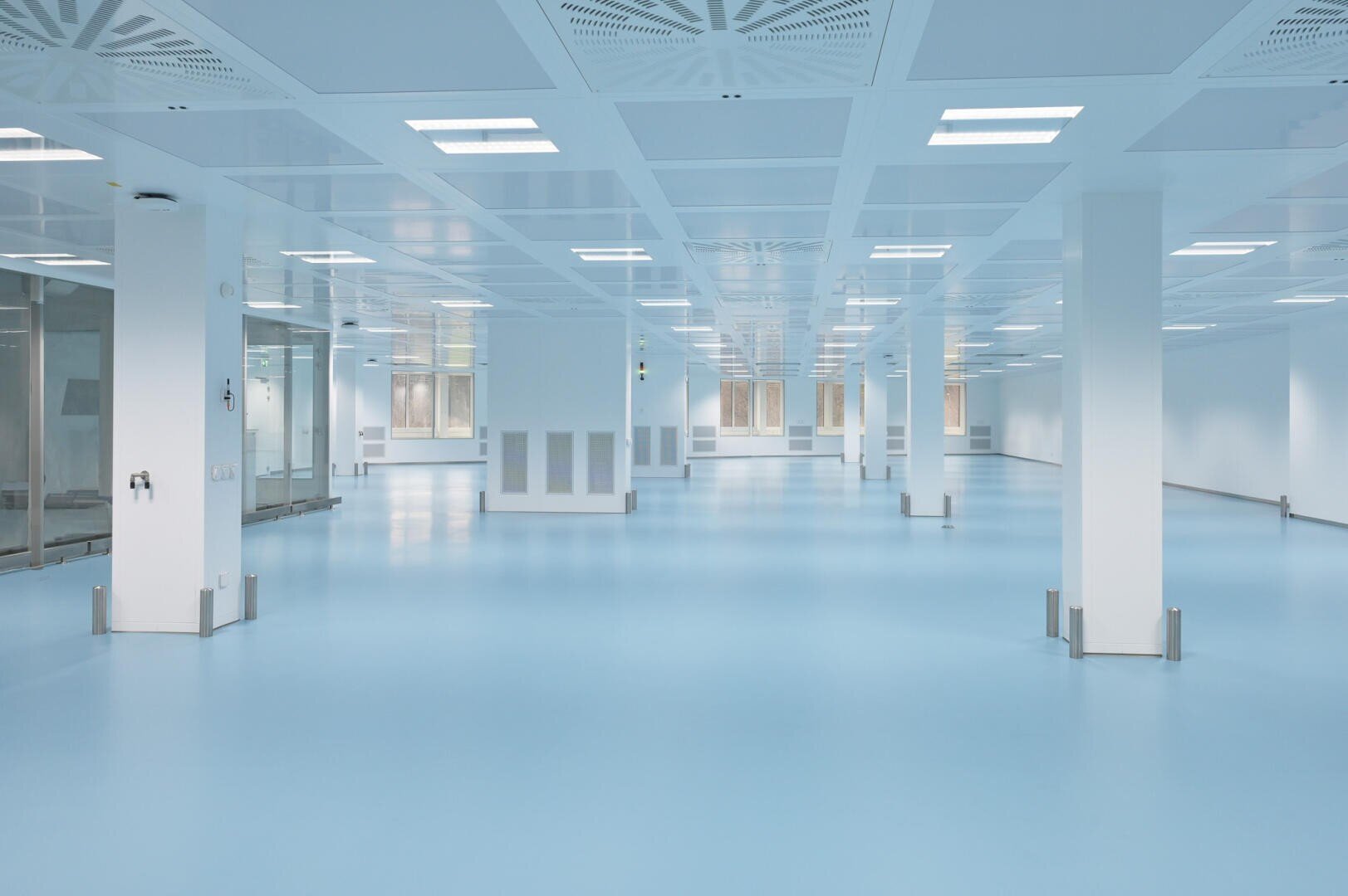 A spacious, empty, modern cleanroom with shiny blue floors, white walls, large ceiling lights and glass doors. The room is brightly lit and uncluttered, with pillars dotted around the room.