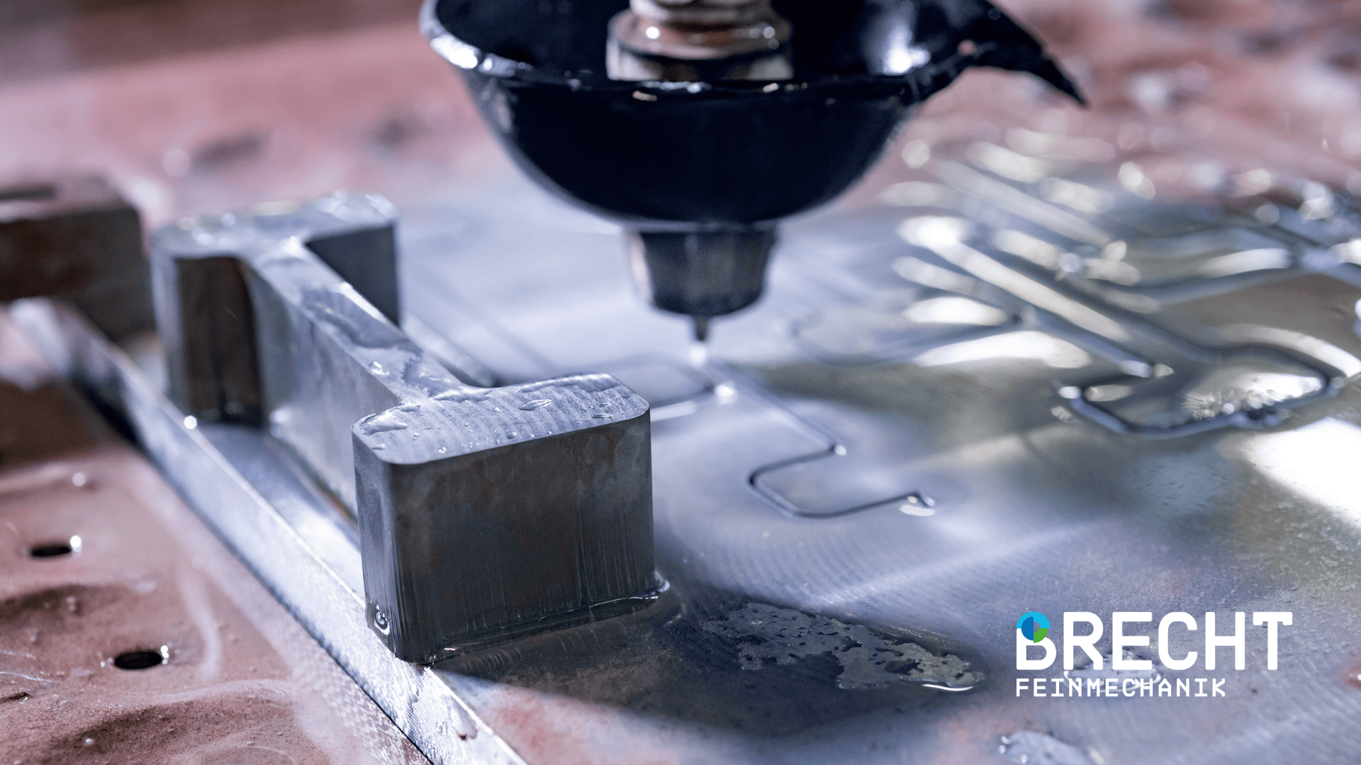 Close-up of a CNC milling machine cutting a metal workpiece with coolant, showing precision engineering. The Brecht Feinmechanik logo can be seen in the bottom right-hand corner.