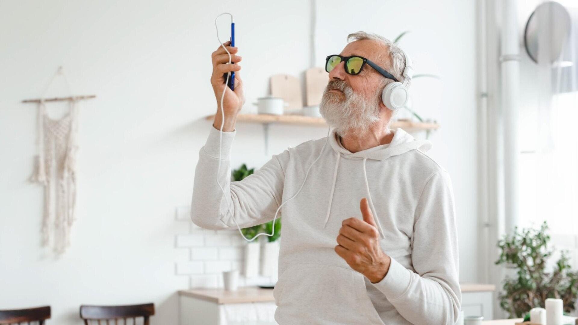 An older man wearing headphones and sunglasses stands in a bright kitchen, holding up a smartphone and smiling as if enjoying music, with one hand raised and wearing a light-colored hoodie.