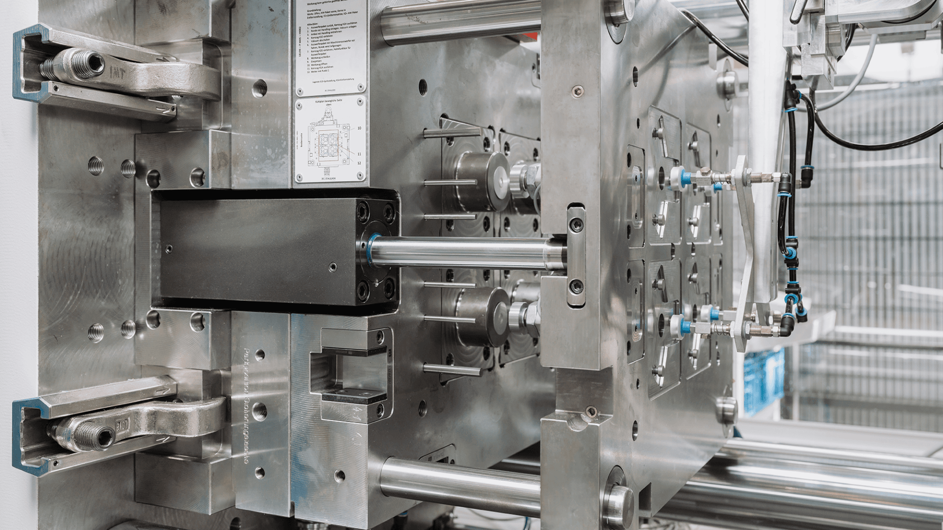 Nahaufnahme einer industriellen Spritzgussmaschine mit einer glänzenden Metallform, Stangen und mechanischen Komponenten, die zur Herstellung von Kunststoffteilen in einer Fabrik verwendet werden.