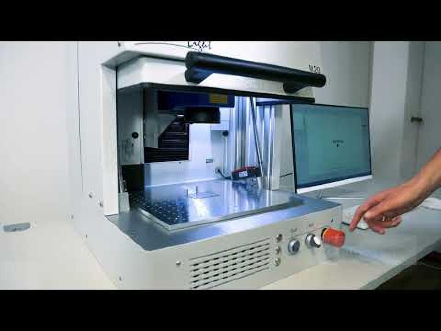 A person's hand presses a red emergency stop button on a desktop laser engraving machine, while a computer monitor can be seen in the background.