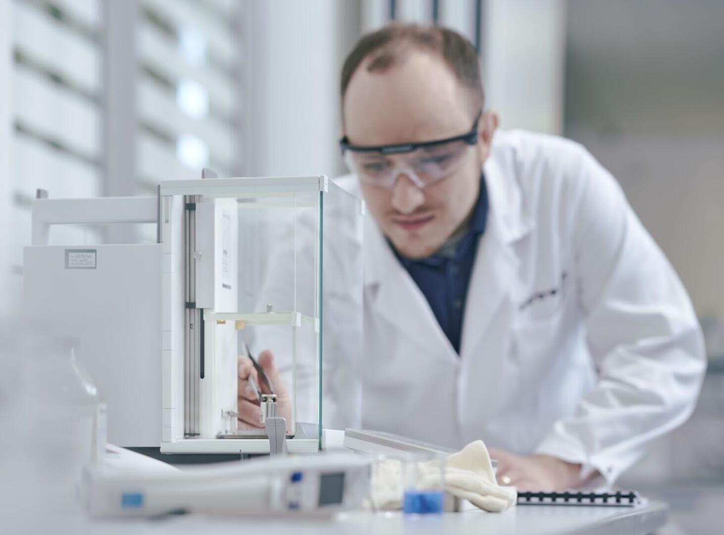 Ein Wissenschaftler in weißem Laborkittel und mit Schutzbrille benutzt in einem Labor sorgfältig eine Präzisionswaage, umgeben von Laborgeräten, darunter ein Notizbuch und Handschuhe.