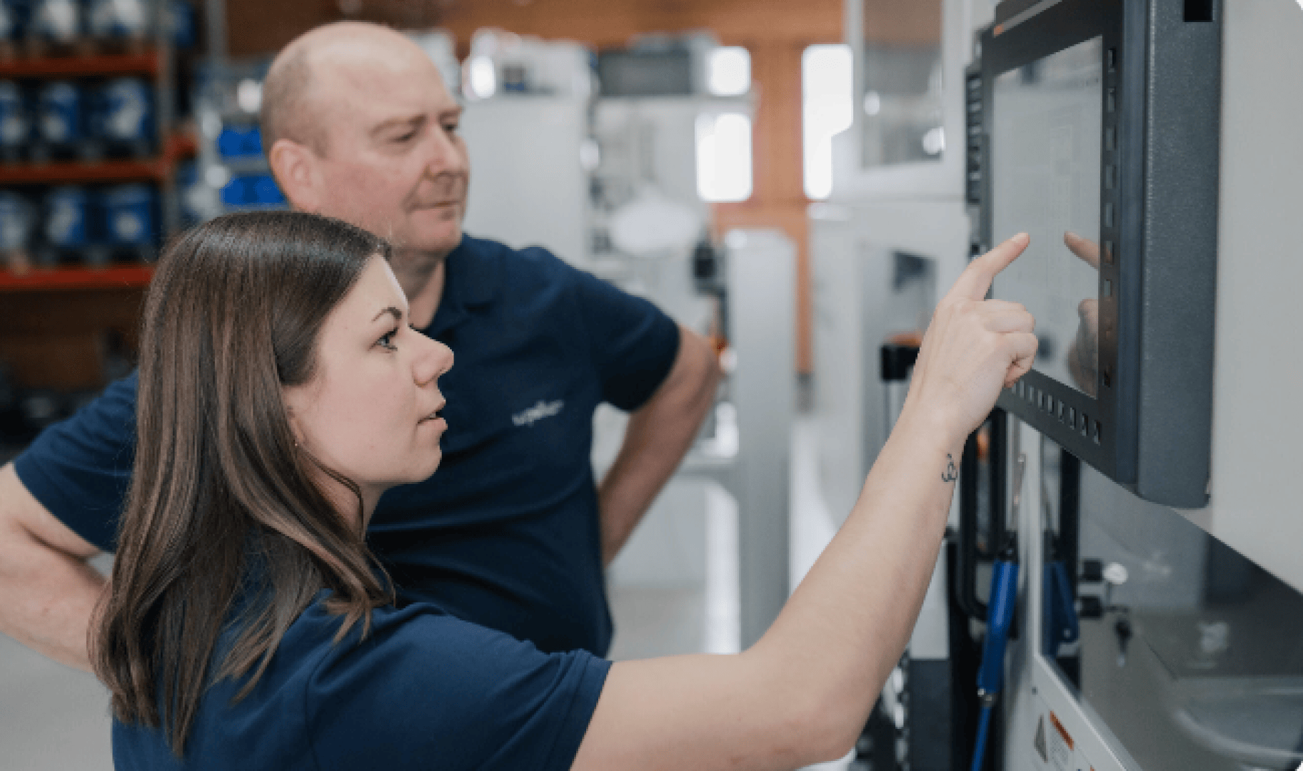 Eine Frau zeigt auf einen Touchscreen an einer großen Maschine, während ein Mann neben ihr steht und beobachtet. Beide tragen marineblaue Hemden in einer Industrie- oder Werkstattumgebung.