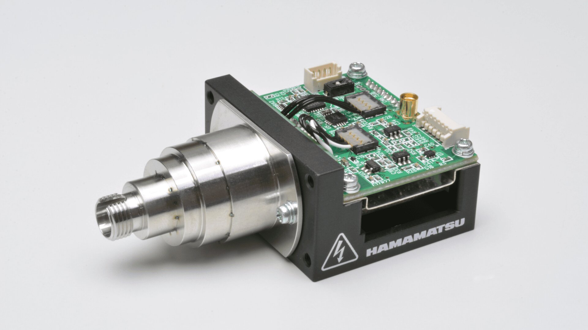 Eine Nahaufnahme eines elektronischen Sensormoduls von Hamamatsu mit freiliegender Leiterplatte, metallischer zylindrischer Komponente und verschiedenen Anschlüssen auf weißem Hintergrund.