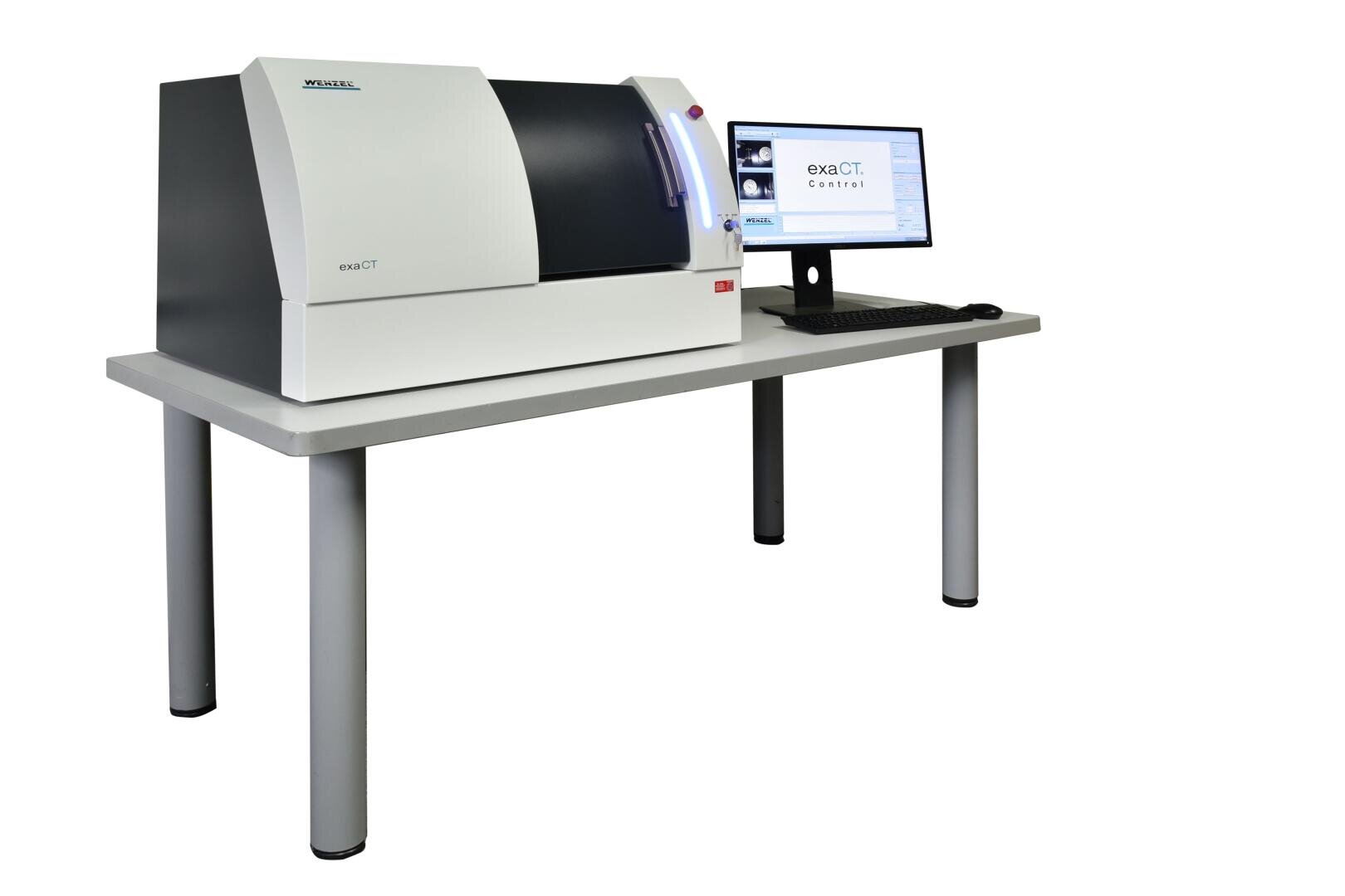 Ein Desktop-CT-Scanner mit weiß-schwarzem Gehäuse steht auf einem Tisch neben einem Computermonitor, auf dem die CT-Scan-Software läuft. Beide stehen auf einem hellen Tisch mit vier Beinen vor weißem Hintergrund.