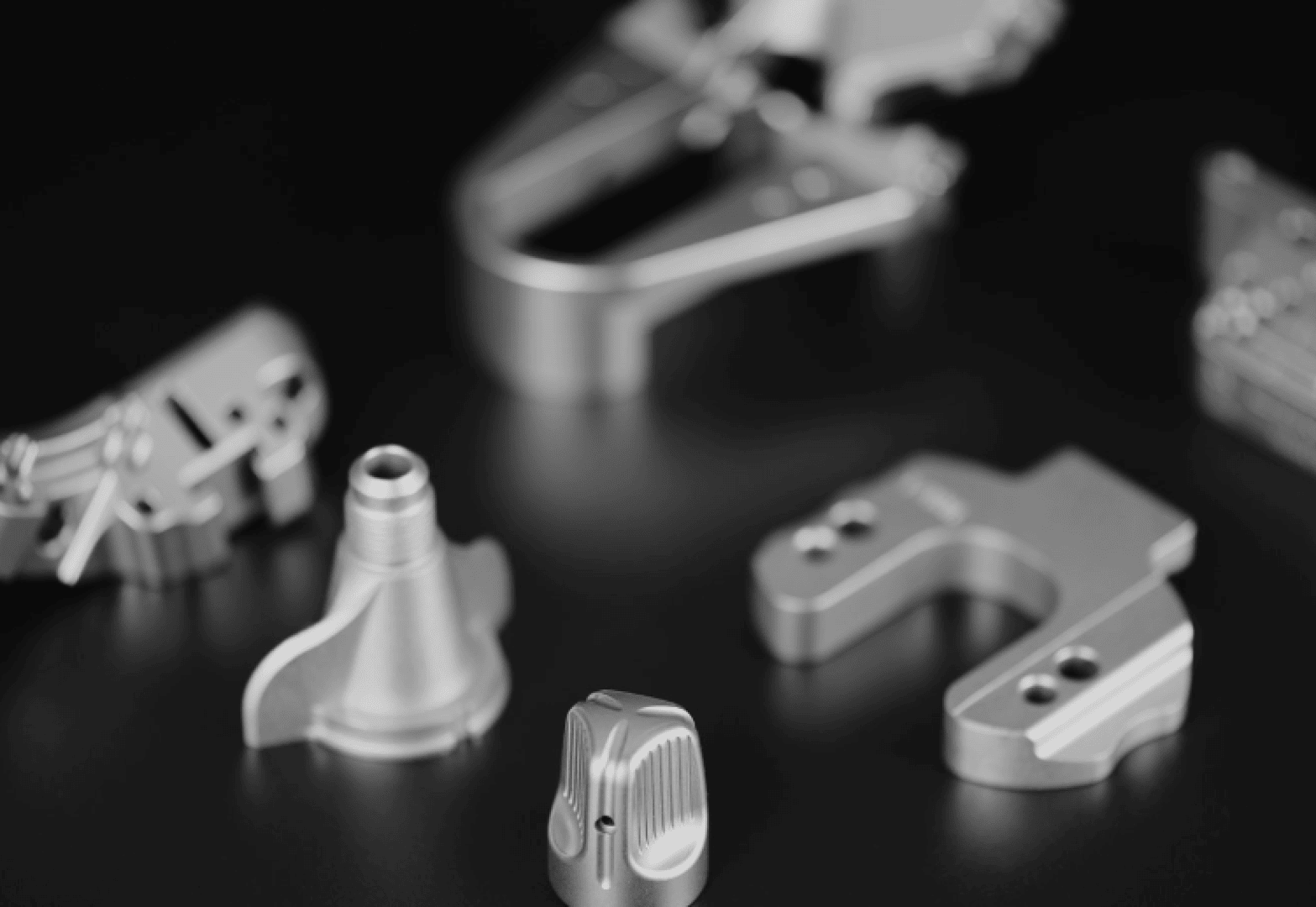 Close-up of various precision-machined metal parts arranged on a black surface, with the central piece in sharp focus and the surrounding components blurred in the background.