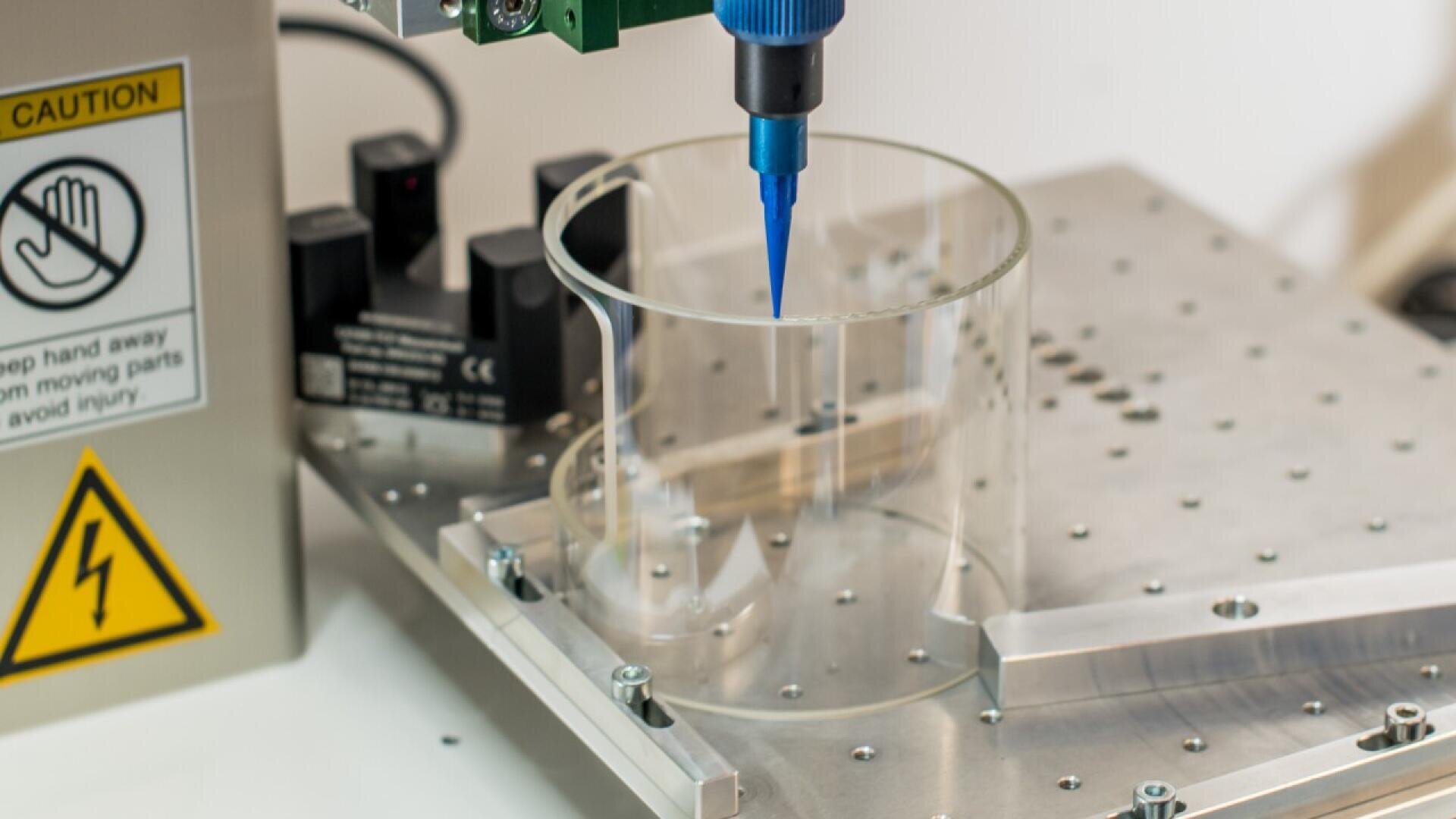 Eine Nahaufnahme einer 3D-Biodruckerdüse über einer transparenten Petrischale auf einer Metallplattform. Auf der Maschine sind Warnschilder zu sehen, die vor beweglichen Teilen und elektrischen Gefahren warnen.
