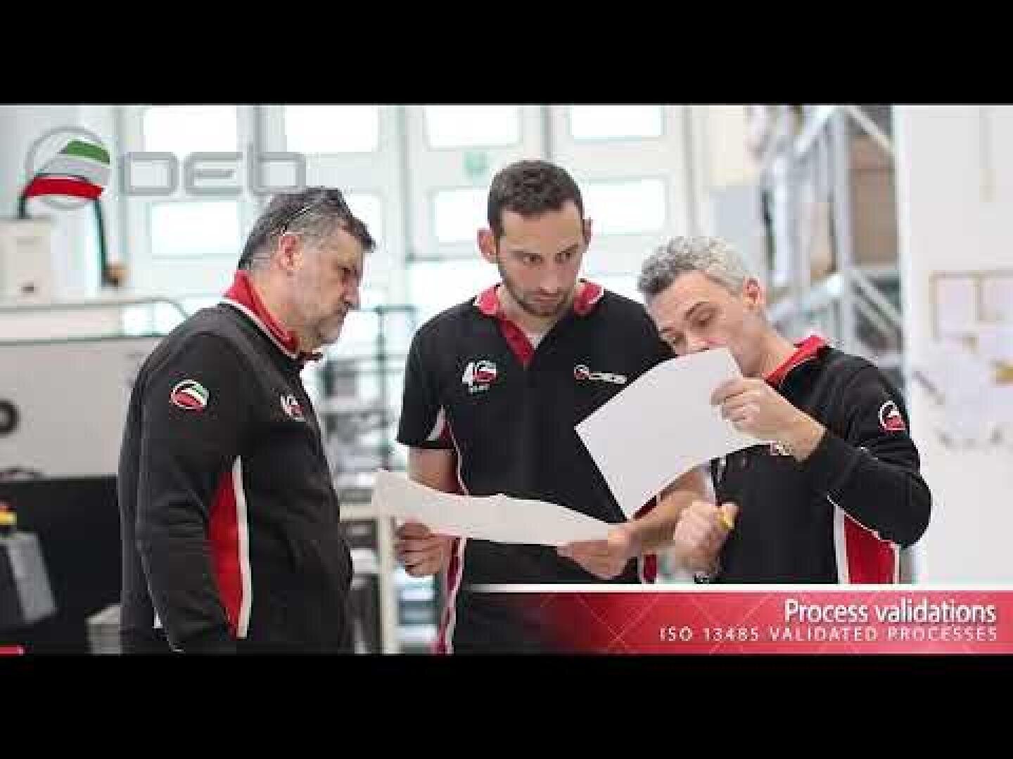 Three men in matching black shirts examine papers together in a bright industrial workspace. The text on the image mentions Process validations and ISO 13485 VALIDATED PROCESSES.