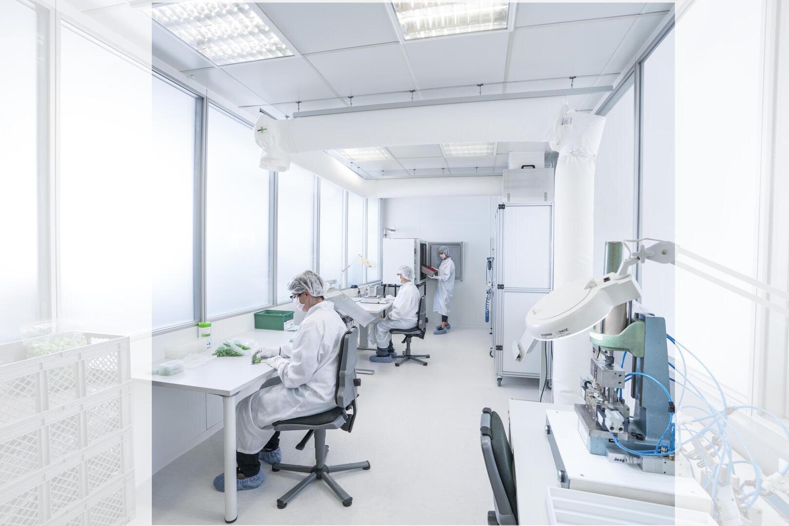 Three people in white lab coats, hairnets and masks work at desks in a bright, modern laboratory filled with scientific equipment and supplies. Large windows provide plenty of natural light.
